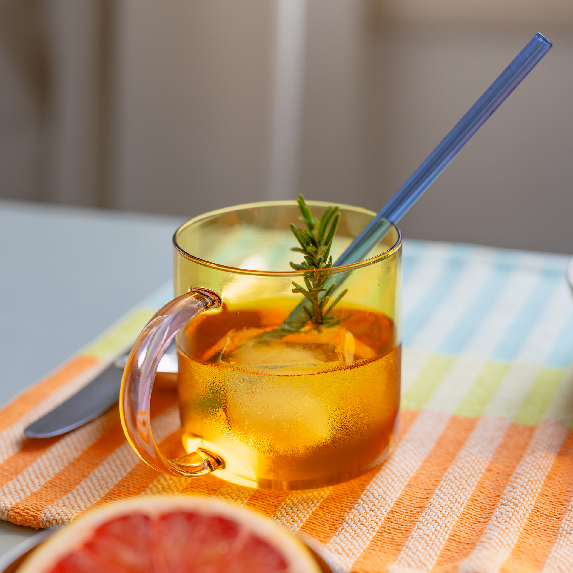 Der Glasbecher „Fred“ in Rosé-Gelb mit Henkel ist mit einem Getränk, Eiswürfeln und einem Zweig Rosmarin befüllt. Ein blauer Strohhalm steckt im Glas. Im Vordergrund ist der Anschnitt einer halben Grapefruit zu sehen.