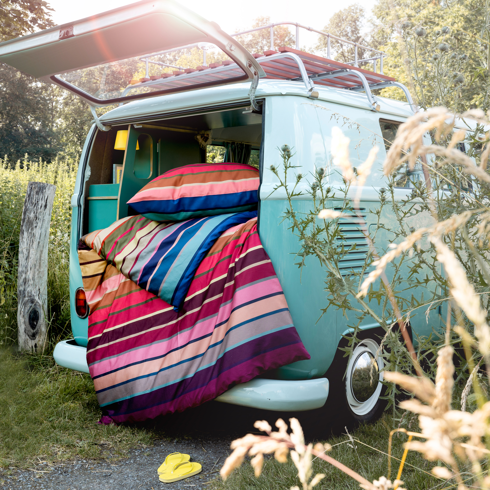 Bettwäsche 'Ascona' im VW-Bus. Bunte, gestreifte Bettwäsche und Kissen liegen im geöffneten Heck eines hellblauen Busses. Draußen stehen gelbe Flip-Flops.