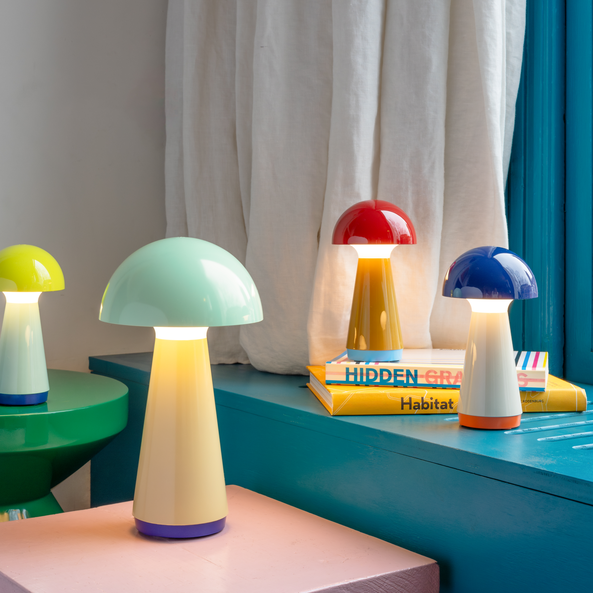 Mehrere 'Bobbi' Tischleuchten in verschiedenen Farben stehen auf Oberflächen und Büchern, einige leuchten. Die Lampen haben eine Pilzform und sind in blau, rot, gelb und grün.