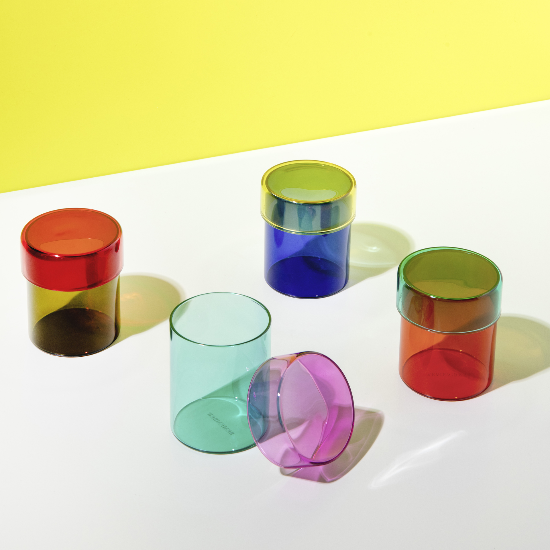 Fünf farbige Glasdosen, eine liegt. Farben sind Rot, Blau, Grün, Türkis und Lila. Sie stehen auf einem weißen Untergrund vor gelbem Hintergrund.