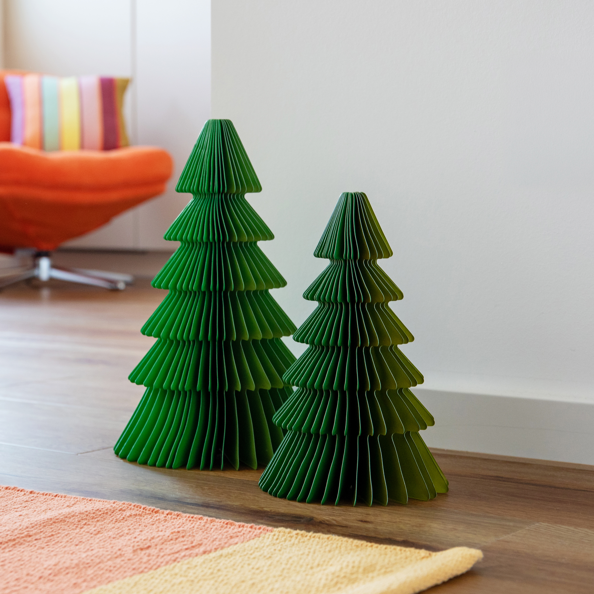 Deux arbres en papier vert plissé sont posés sur un plancher en bois, l'un grand, l'autre petit. Leurs silhouettes en gradins rappellent des sapins stylisés. Le vert bicolore les rend modernes et festifs. Au fond, un fauteuil orange.