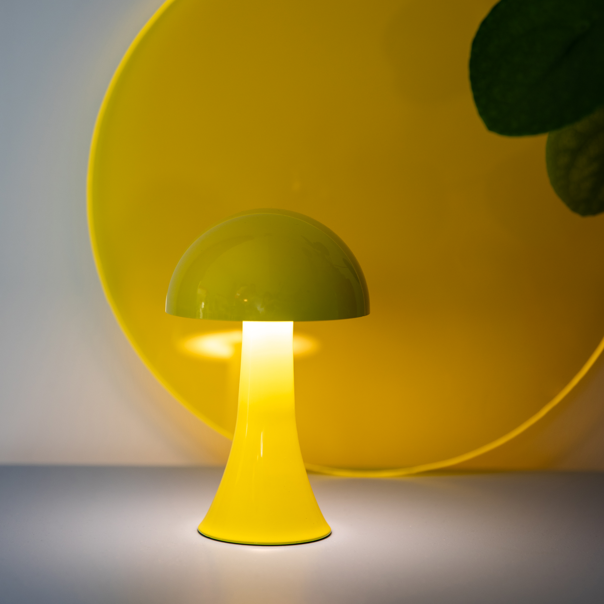 Eine gelb-grüne Lampe im Pilzdesign leuchtet auf einem grauen Tisch vor einem gelben Hintergrund mit Blattwerk.