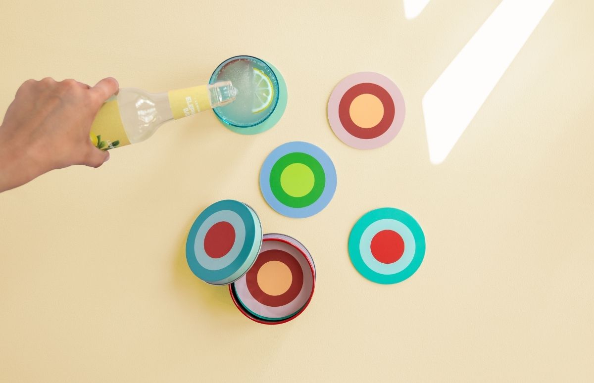Ein Produktbild zeigt eine Hand, die Limonade aus einer gelben Flasche in ein türkisblaues Glas mit Eiswürfeln und Zitronenscheibe gießt. Im Hintergrund liegen runde Untersetzer mit bunten Zielscheibenmustern als Cross-Selling-Artikel.