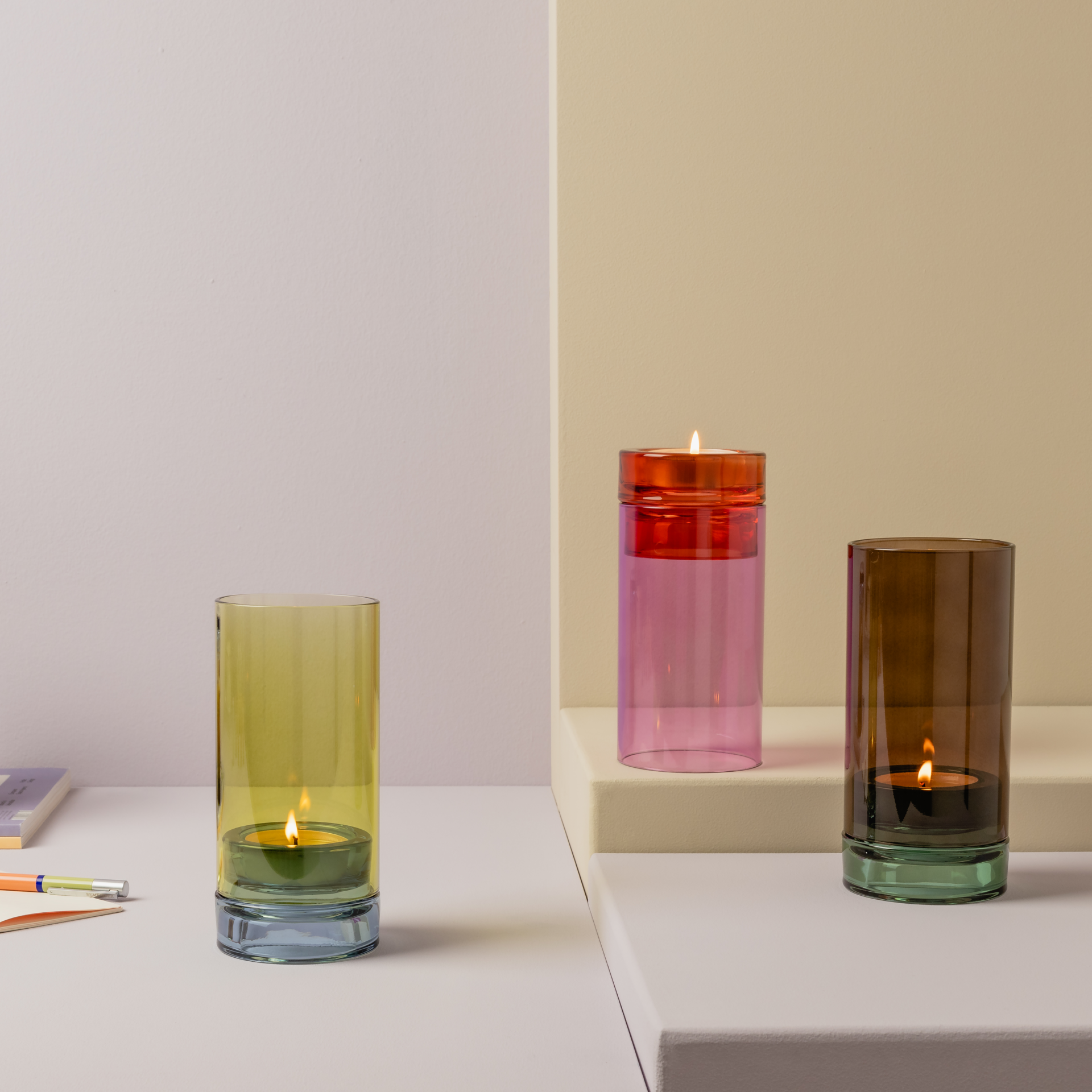 Drei bunte 'Lys' Windlichter aus Glas mit Teelichtern in Gelb, Pink und Braun stehen auf einem weißen Sockel vor pastellfarbenem Hintergrund.