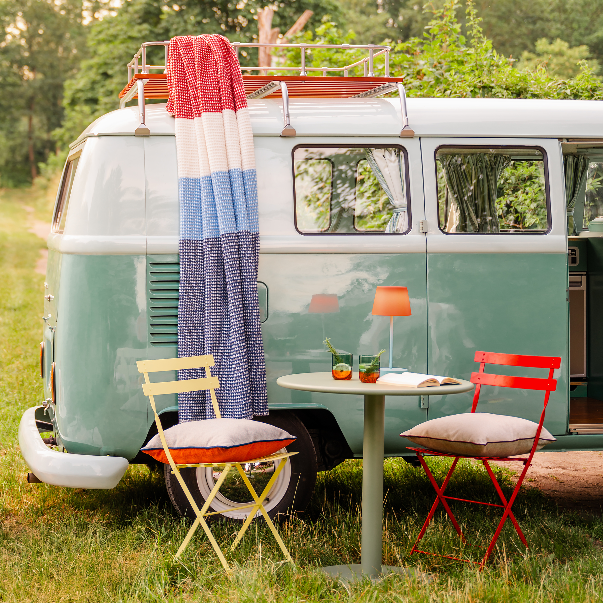 Ein mintfarbener VW-Bus mit rot-weiß-blauer Decke 'Malie' auf dem Dachgepäckträger. Davor ein Tisch mit Lampe und zwei Stühle mit Kissen im Grünen.