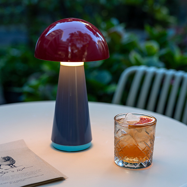 Moderne pilzförmige Tischlampe mit dunkelrotem Schirm und blau-türkisem Farbverlauf am Fuß auf hellem Außentisch. Sie leuchtet sanft und beleuchtet ein Glas mit Eis, Orangendrink und Grapefruitscheibe.