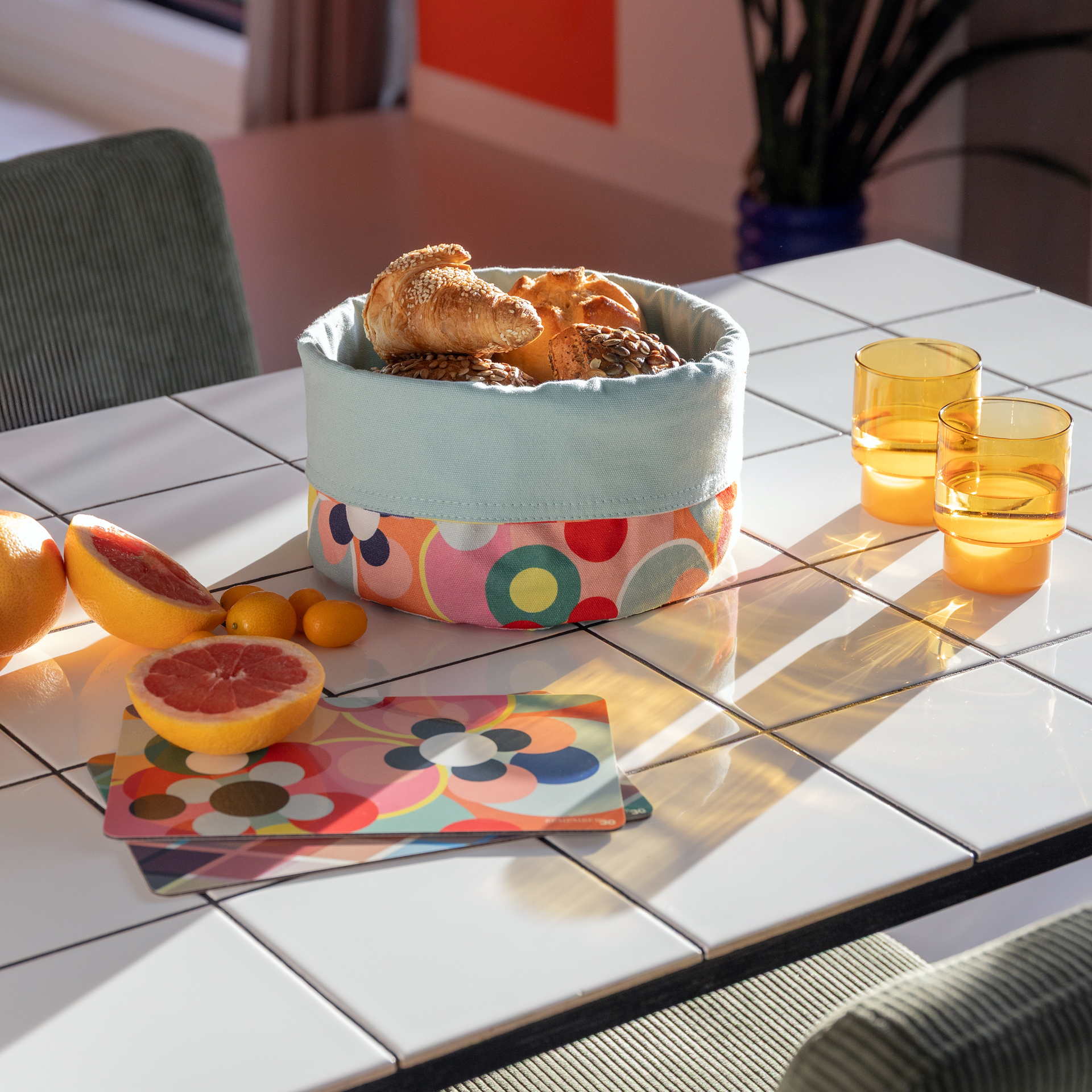 Ein Tisch mit weißer Fliesenoberfläche präsentiert einen Korb mit Gebäck, Grapefruit, Kumquats, zwei gelben Gläsern und ein Schneidebrett mit Blumenmuster. Sonnenschein akzentuiert die Szene.