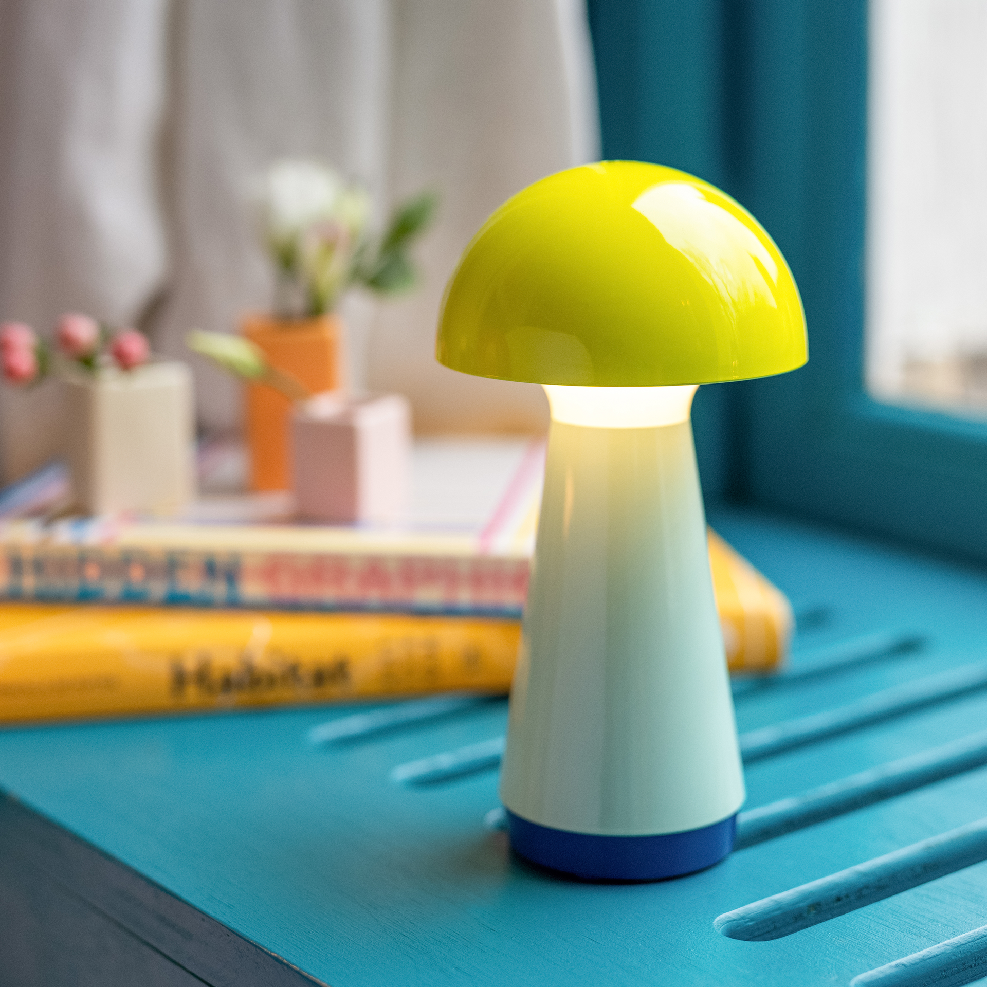 Gelbe Tischleuchte Bobbi im Ambiente. Die Leuchte hat eine Pilzform und spendet warmweißes Licht. Im Hintergrund sind Bücher und Dekorationen zu sehen.