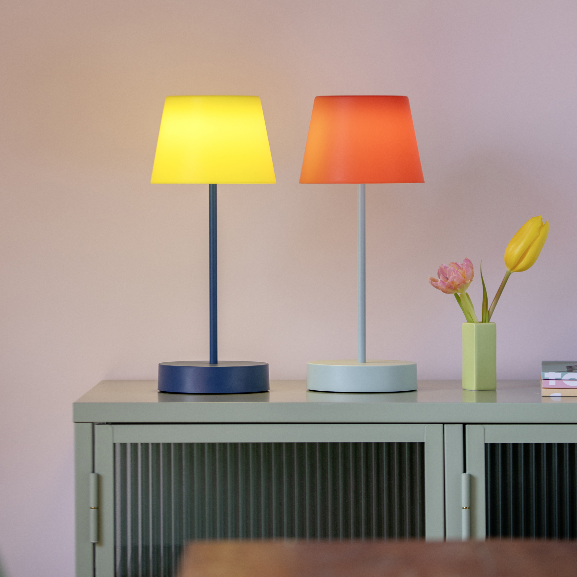 Zwei Tischleuchten 'glory' in Gelb und Orange auf einem Sideboard neben Blumen in einer Vase vor einer rosa Wand.