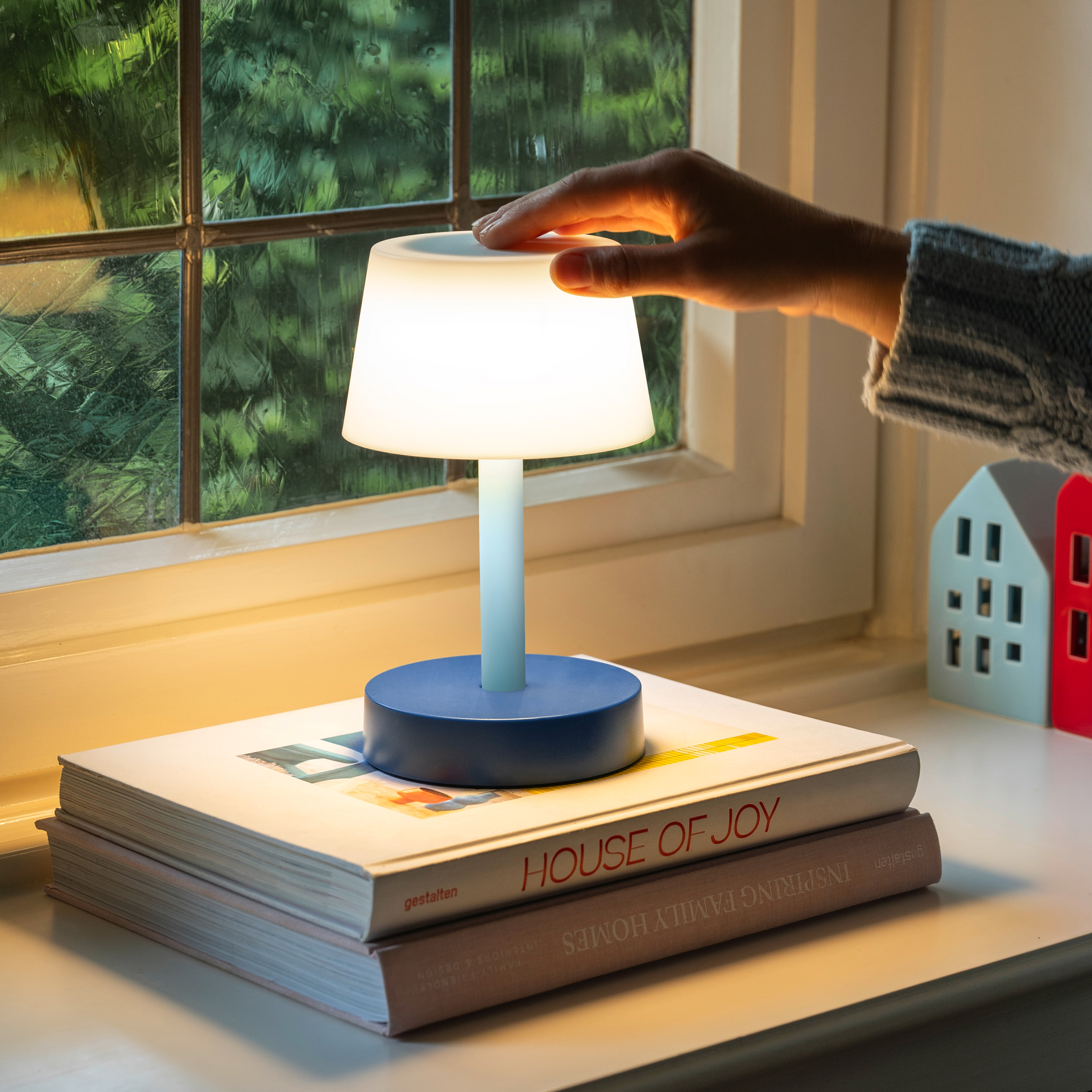 Das Produktbild zeigt die Tischleuchte Fritz 'sky' auf Büchern. Eine Hand berührt den leuchtenden Schirm. Im Hintergrund sind ein Fenster und Deko-Häuser zu sehen.