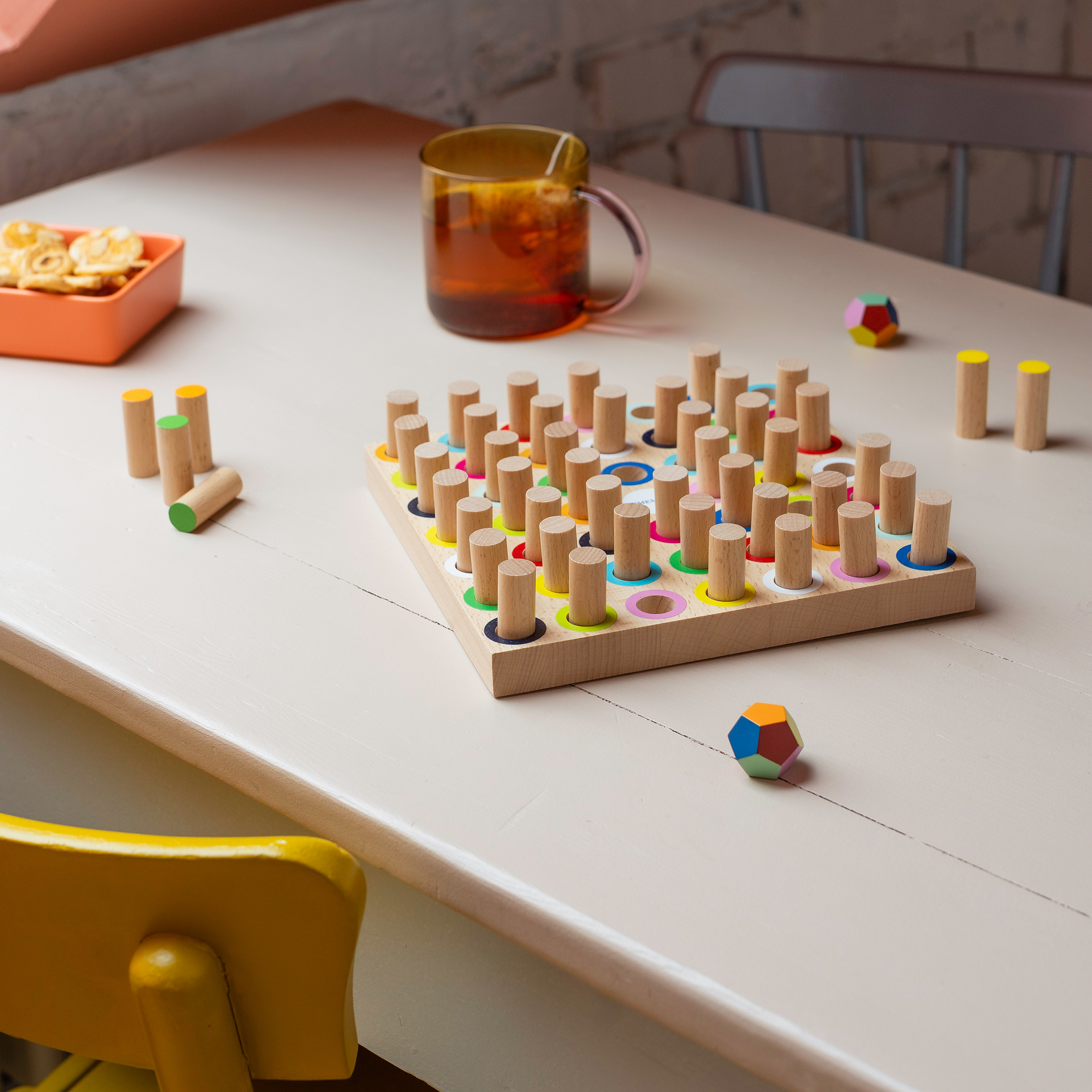 Auf einem Tisch stehen ein Holzspiel mit bunten Ringen, eine Tasse Tee, ein Schälchen mit Snacks und ein bunter Ball. Holzstifte liegen verstreut. Eine gelbe Stuhllehne ist sichtbar.