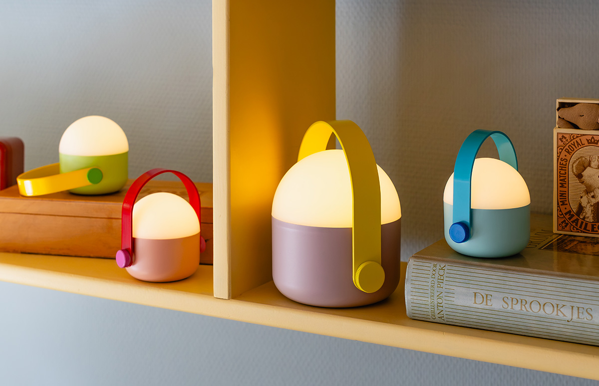 Trois lanternes portables pastel sont sur une étagère jaune clair. À gauche, une petite lanterne verte sur une caisse en bois, près d'une plus grande lavande. À droite, une lanterne bleu clair à poignée turquoise sur un livre.