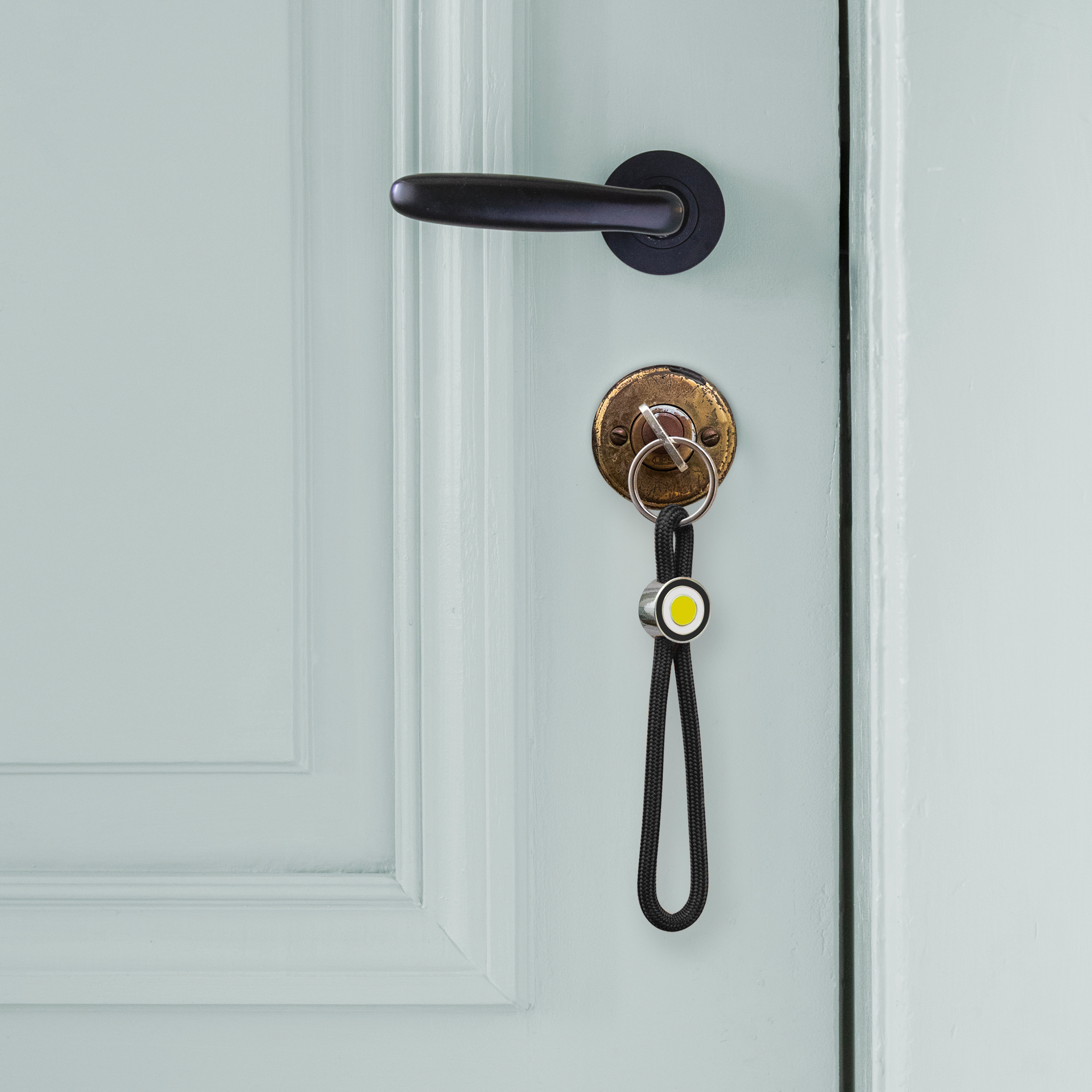 Porte-clés noir en corde tressée, en forme de boucle. Au centre, un anneau métallique brillant maintient la corde, avec un liseré noir et un cercle jaune vif au design Ringo. En bas, un anneau à clés argenté.