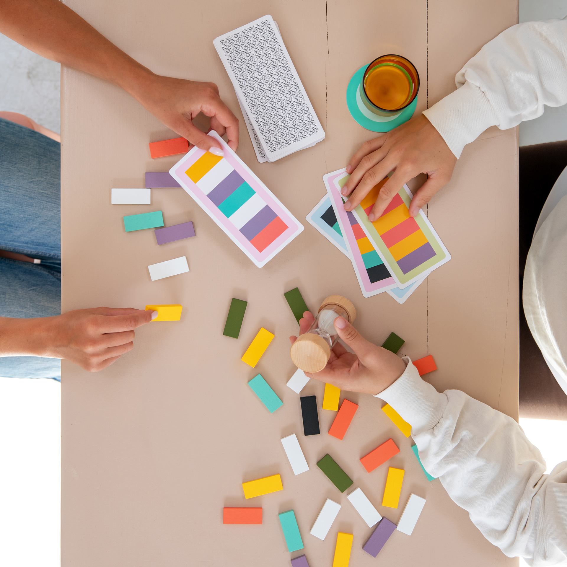 Éléments d'un jeu de mémoire posés sur une table blanche. Des tuiles rectangulaires en bois colorées sont disposées devant une carte de mission présentant le même motif de couleurs. À côté, un sablier presque vide ; à l'arrière-plan, des pions flous.