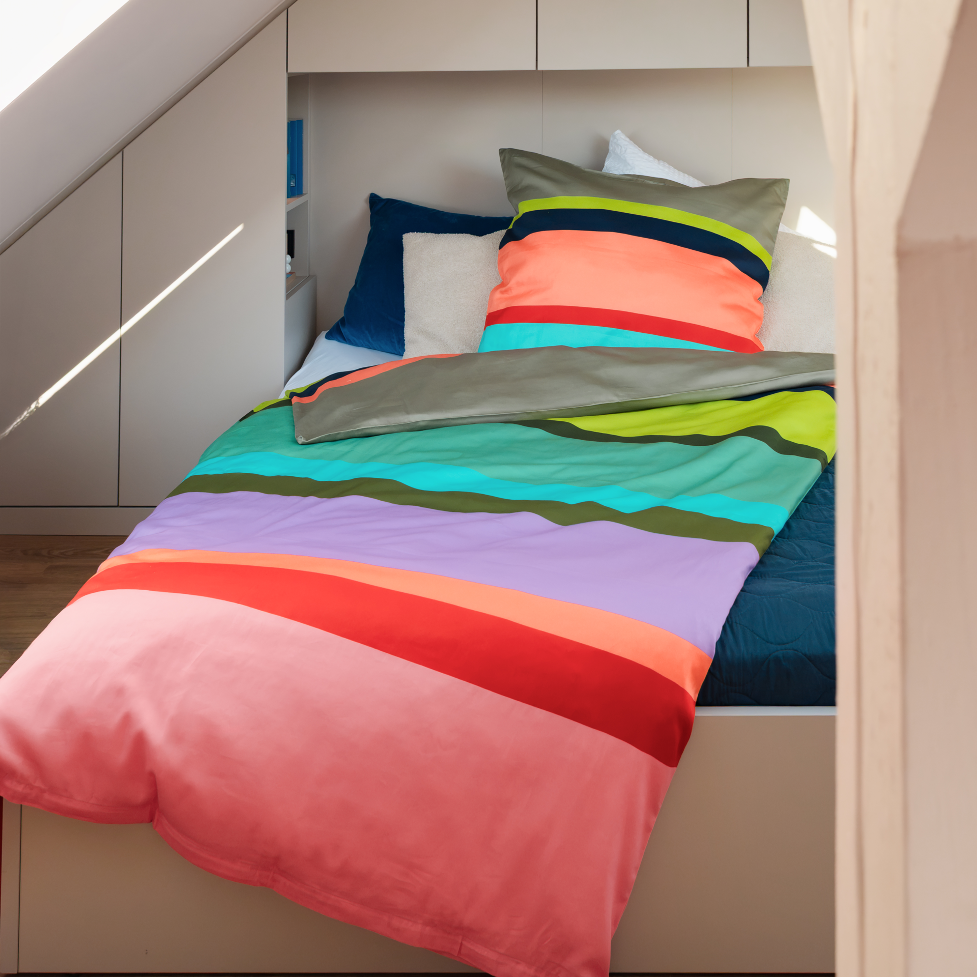 Gefaltete Bettwäschegarnitur aus seidenweichem Mako-Satin mit Kissen- und Bettbezug. Beide Bezüge zeigen breite horizontale Streifen in leuchtenden Farben wie Beige, Gelb, Dunkelblau, Koralle, Rot, Hellblau, Rosa, Lila, Oliv- und Mintgrün.