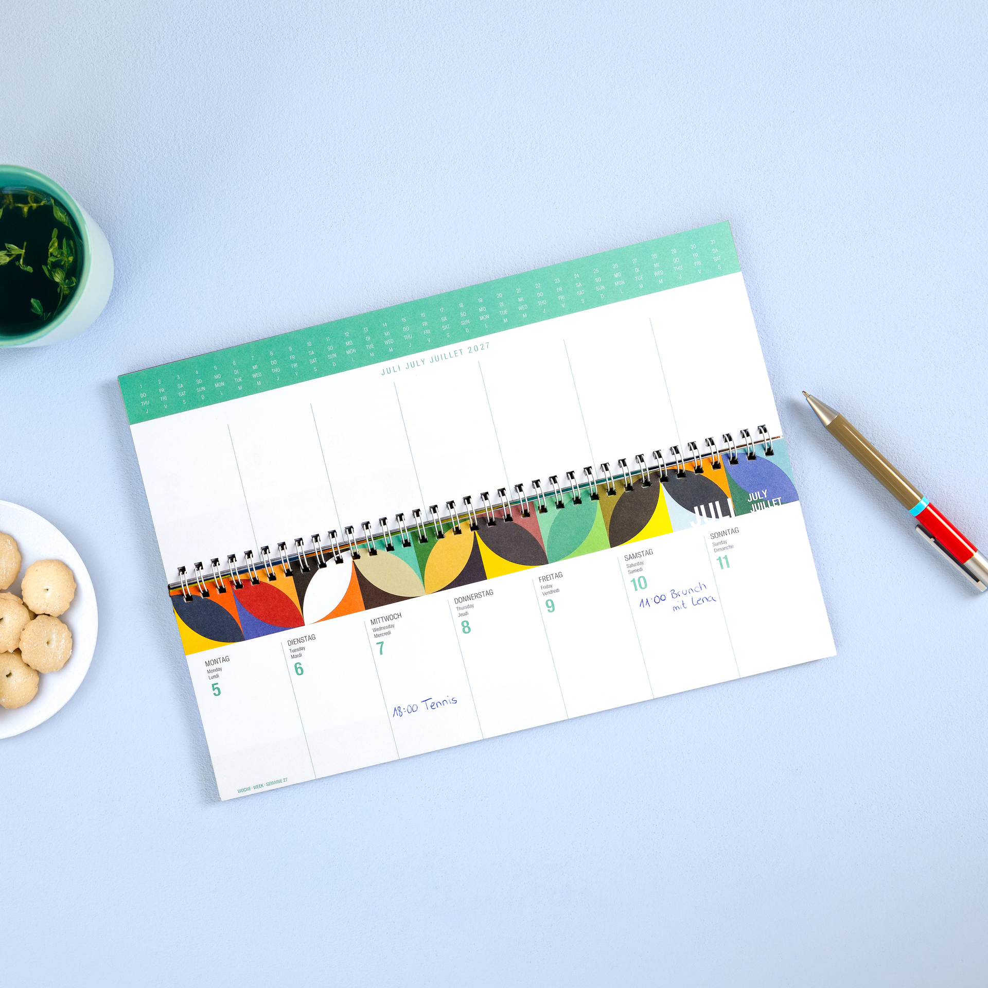 Agenda hebdomadaire coloré 2027 à reliure spirale en haut, idéal pour gérer vos rendez-vous sur votre bureau. Couverture : motif abstrait de formes géométriques bleues, vertes, rouges et jaunes. Texte blanc : «REMEMBER» et «AGENDA HEBDOMADAIRE 2027».