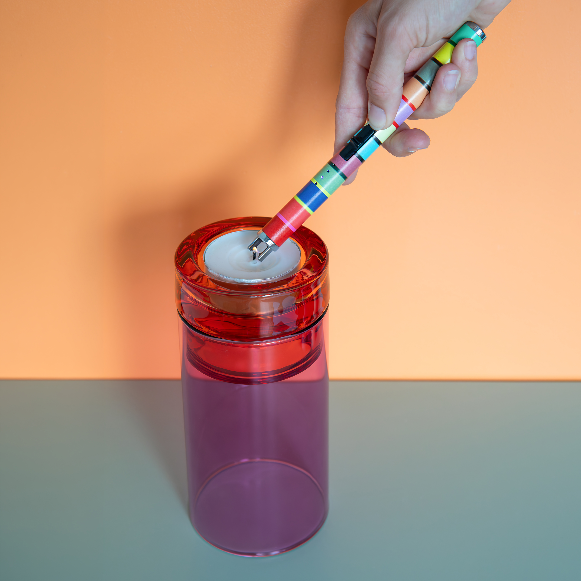 Una mano regge un accendino a bastoncino sottile, a strisce colorate, che accende una piccola candela da tè bianca in un bicchiere rosso. L'accendino elettrico dal design " REMEMBER" produce un arco elettrico.