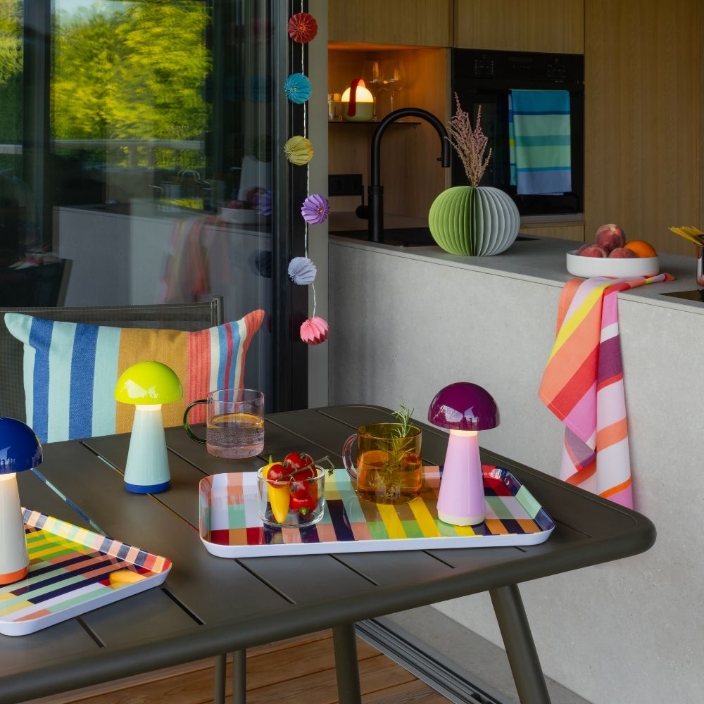 Terrasse extérieure moderne avec table foncée et objets colorés. Trois lampes champignon bleue, vert clair et violette, près de plateaux rayés avec boissons et en-cas. Cuisine lumineuse, fenêtres sur les arbres. Guirlandes en papier.