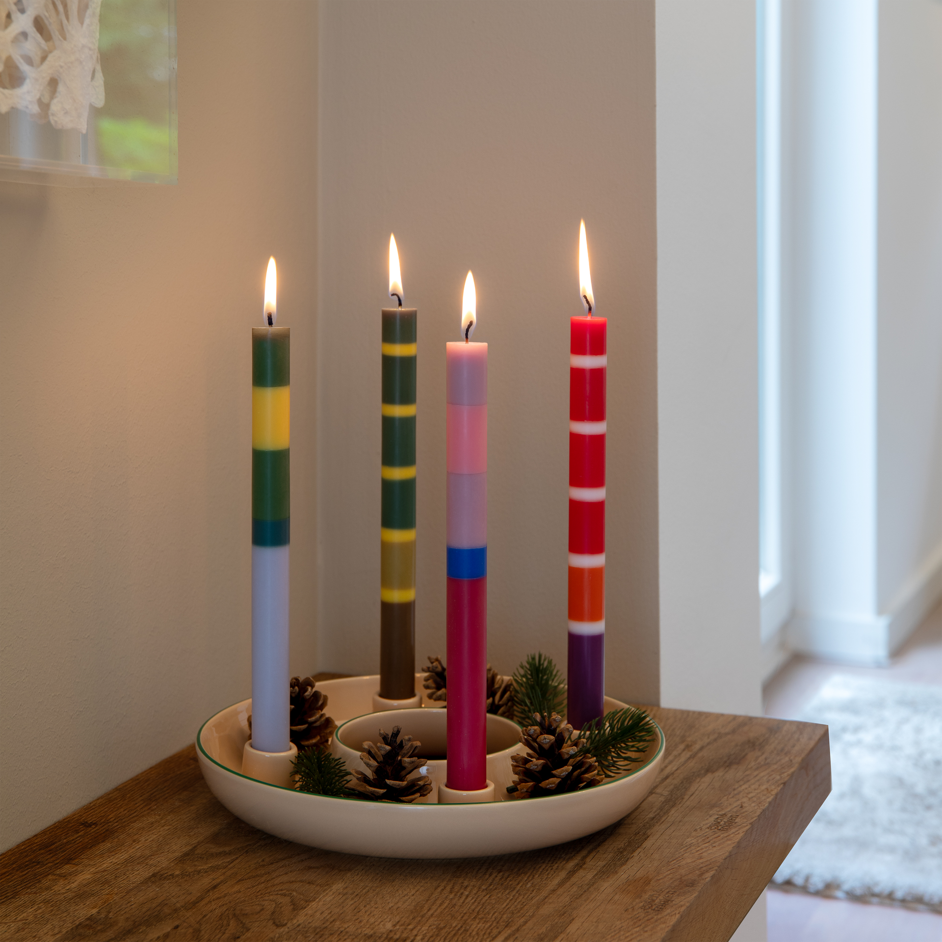 Su un tavolo di legno è posato un portacandele beige con il bordo verde. Al suo interno bruciano quattro candele a bastoncino con strisce colorate nei toni del verde, giallo, blu, rosa e rosso. Tra le candele sono disposte pigne e rametti verdi.