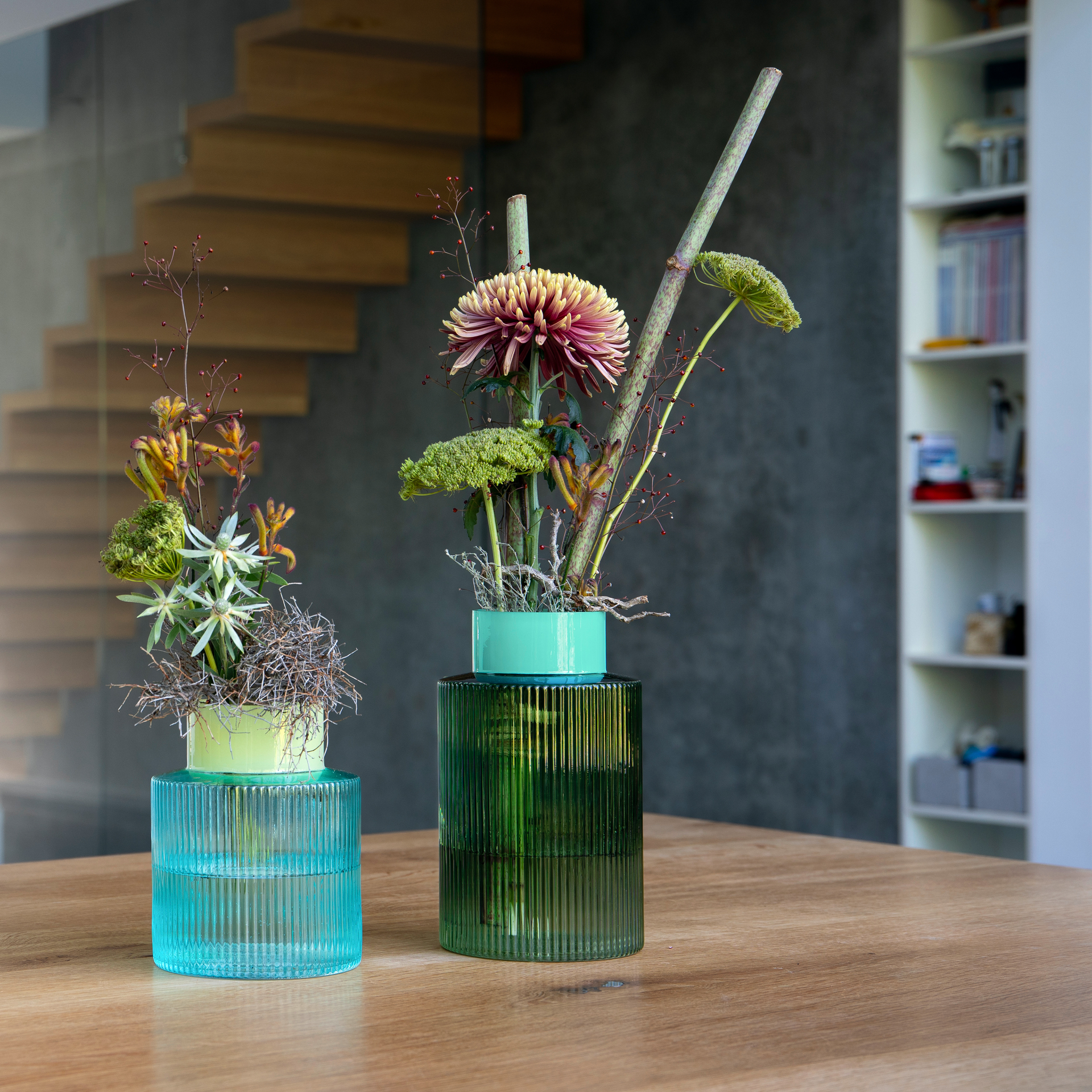 Zwei geriffelte Glasvasen, eine grüne, eine blaue, mit floralen Dekorationen auf einem Holztisch. Im Hintergrund Treppe und Regal.