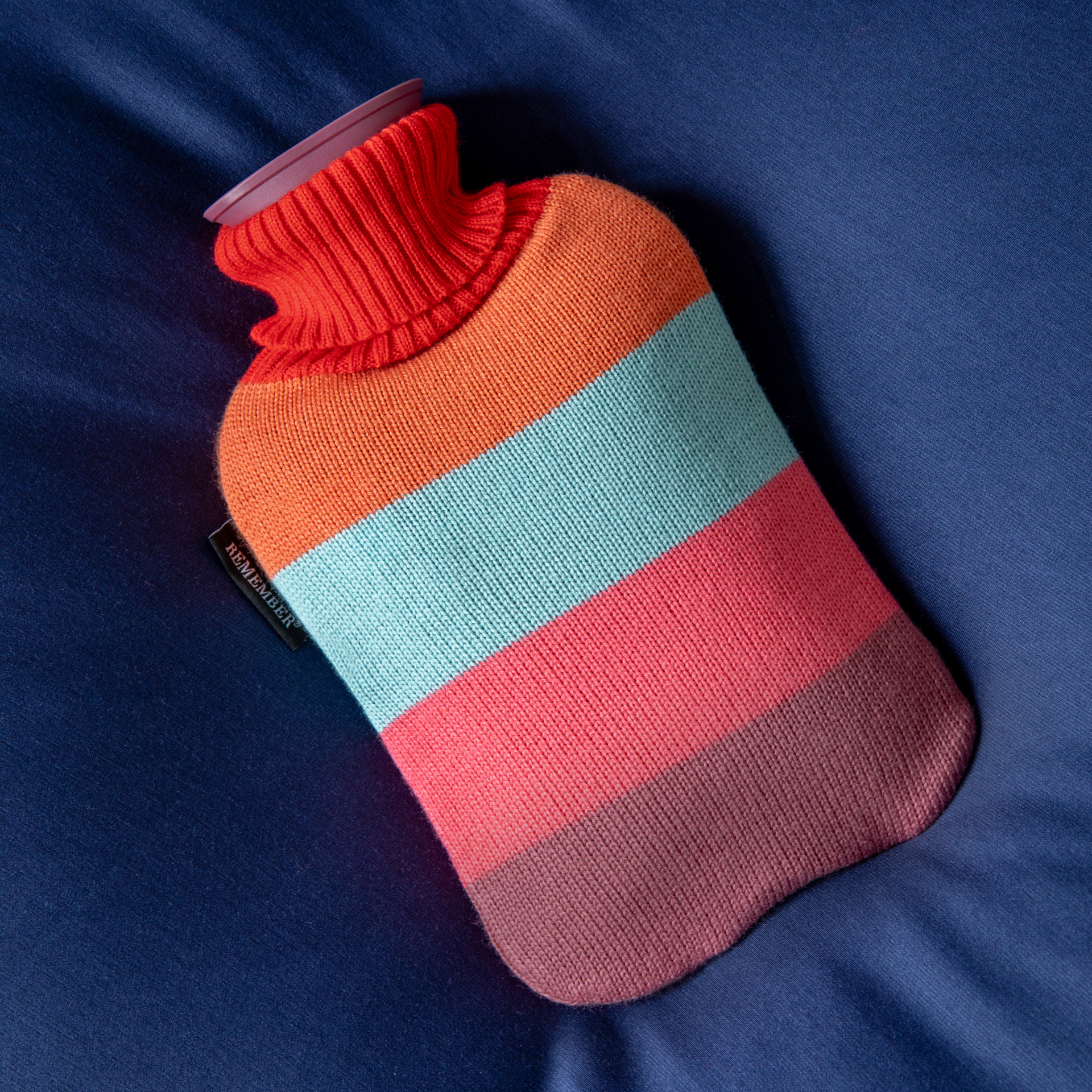 Wärmflasche 'Lilli' mit farbigen Streifen (orange, blau, rosa und braun) auf blauem Stoff. Der Verschluss ist lila.