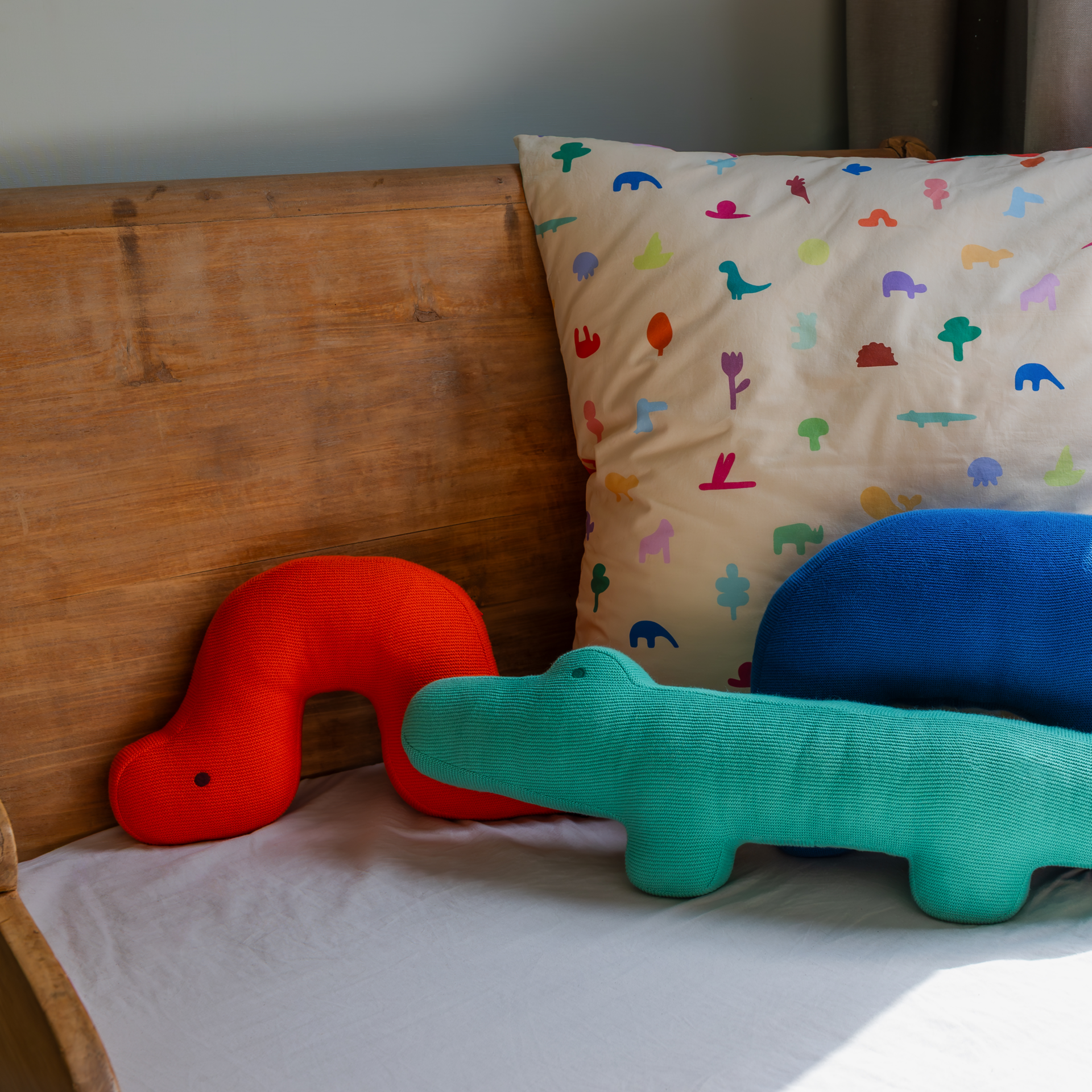 Kuschelkissen 'Raupe Frieda' und weitere Kissen auf einem Bett. Die Raupe ist rot, daneben ein blaues und ein grünes Kissen. Das Rückenkissen ist mit bunten Motiven bedruckt.