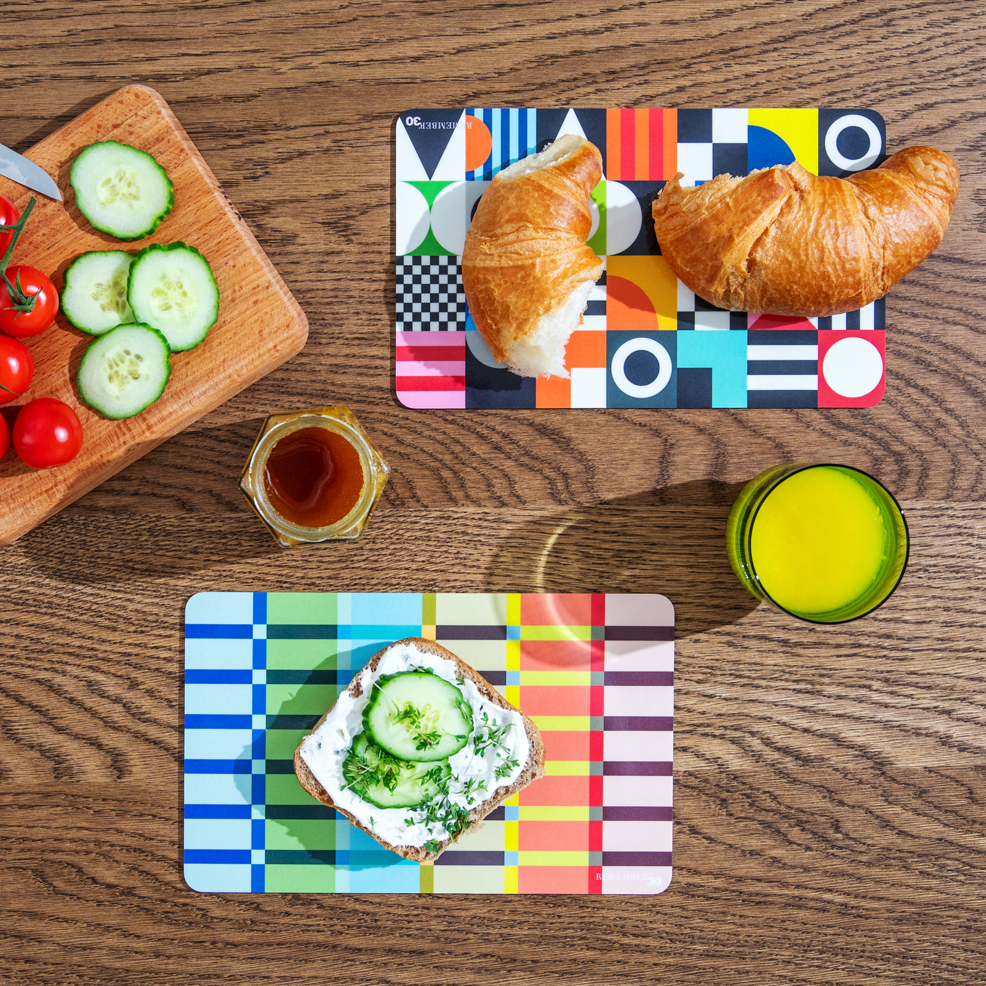 Vista dall'alto di un tavolo per la colazione con croissant, pane con crema di formaggio e cetrioli, pomodori, miele e succo di frutta su tovagliette colorate.