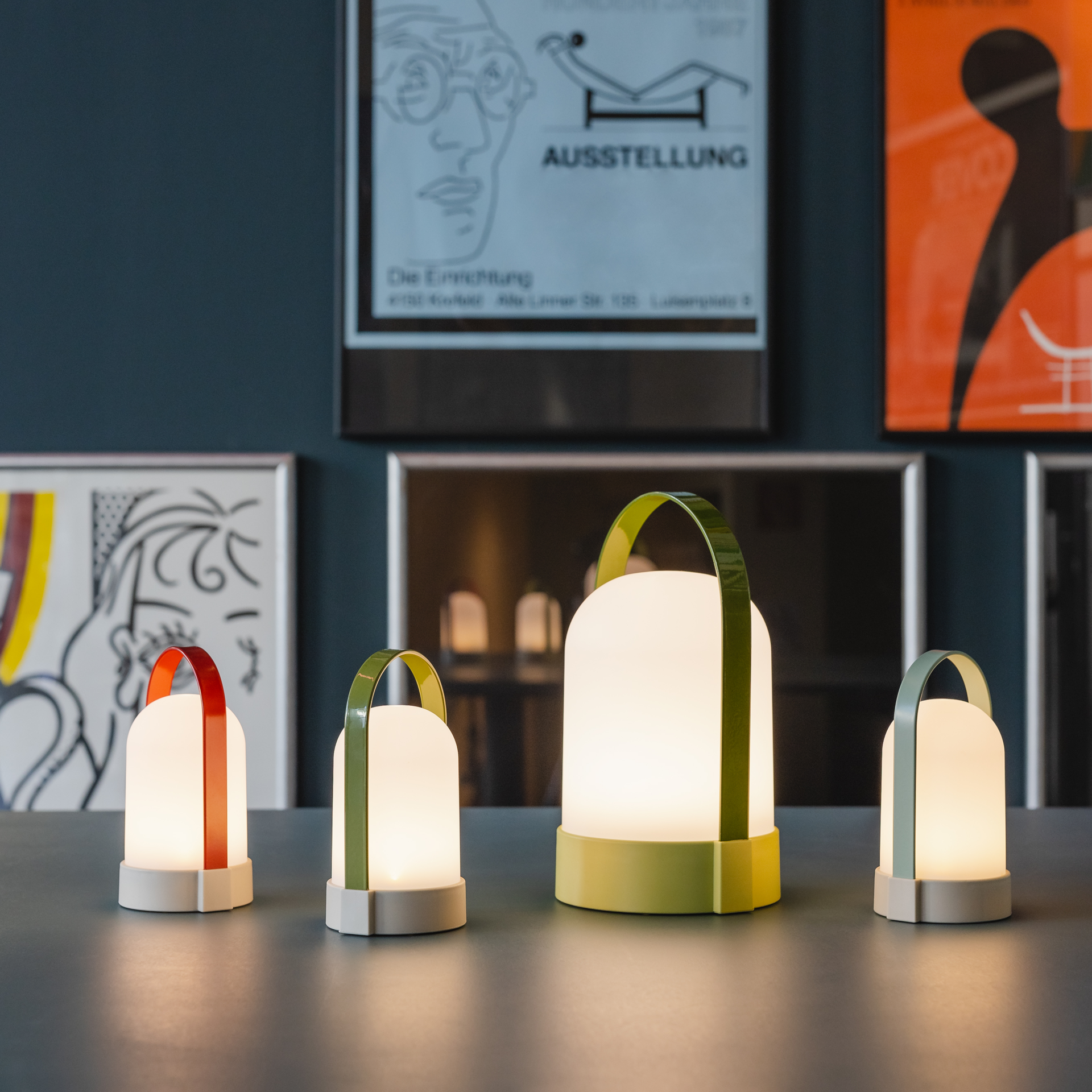 Drei URI Piccolo Leuchten im Set mit farbigen Tragebügeln in Rot, Hellblau und Grün auf einem Tisch. Im Hintergrund sind Kunstdrucke an einer Wand zu sehen. Die Leuchten sind eingeschaltet und strahlen warmweißes Licht aus.