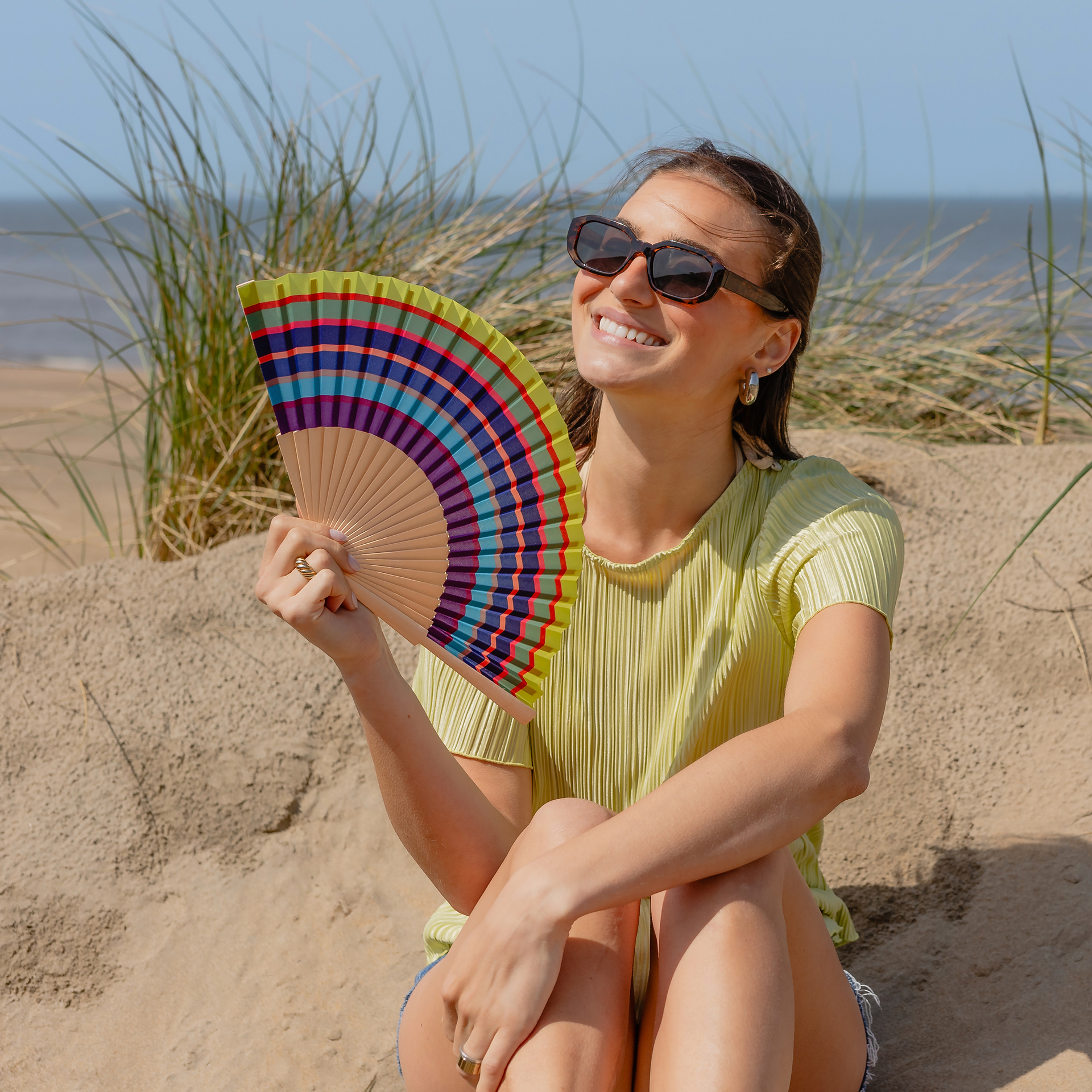 Frau nutzt am Strand den 'Costa' Handfächer als persönliches Ventilationsgerät. Der Fächer weist ein buntes Striped-Design auf. Sie trägt eine Sonnenbrille und lacht. Dünengras im Hintergrund.