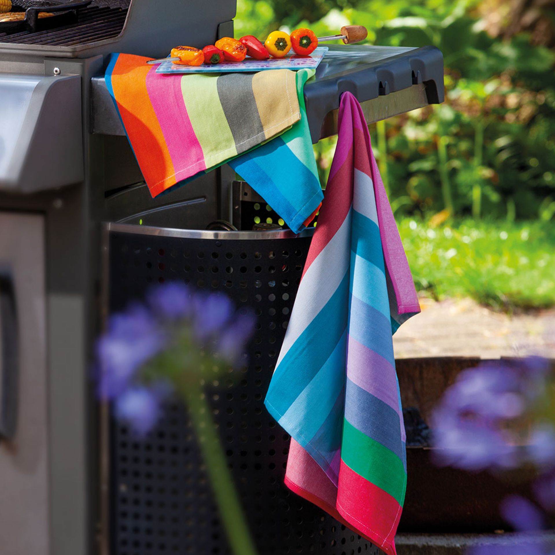 Ein Trockentücher-Set mit bunten Streifen, drapiert auf einem Grill. Auf einem der Tücher liegen gegrillte Paprika.