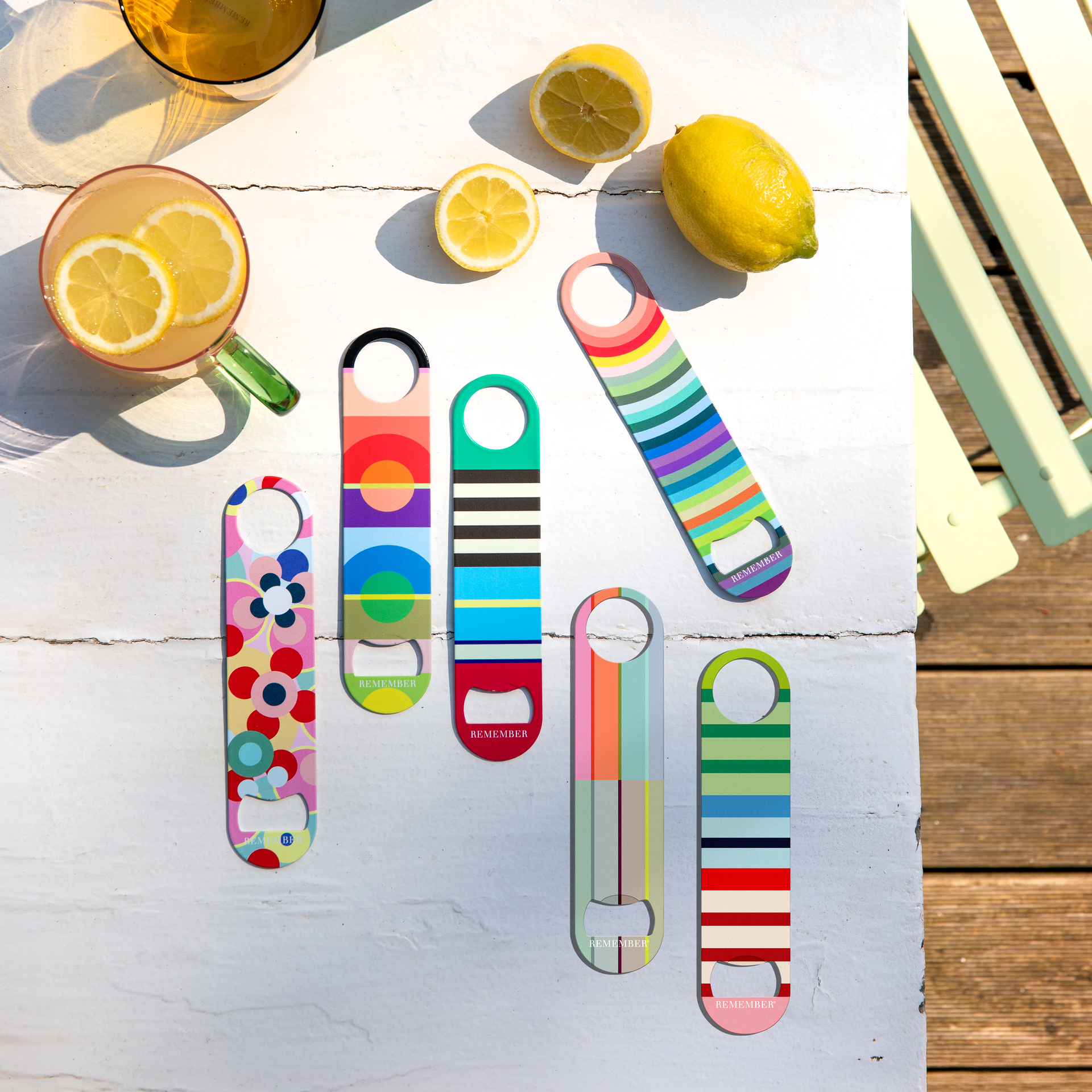 Sechs bunte Flaschenöffner mit auffälligen Mustern liegen in der Sonne auf einem weißen Holztisch. Dazu zwei Gläser Limonade mit Zitronenscheiben sowie ganze und halbierte Zitronen. Im Hintergrund ein hellgrüner Holzstuhl.