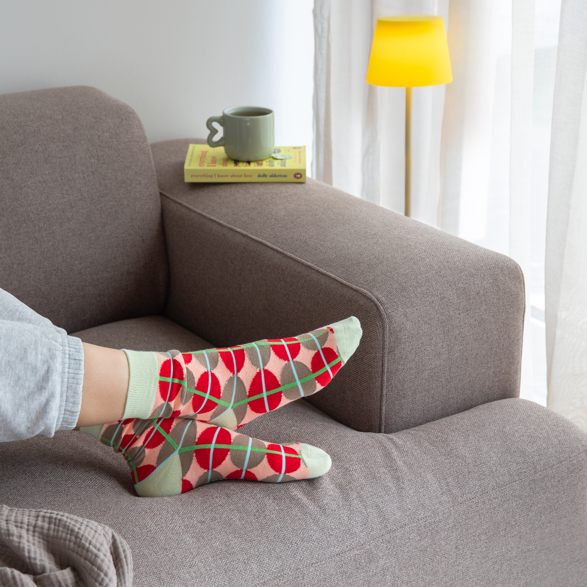 Person mit gemusterten Socken auf braunem Sofa. Buch und Tasse stehen auf der Sofalehne. Gelbe Lampe und Vorhang im Hintergrund.