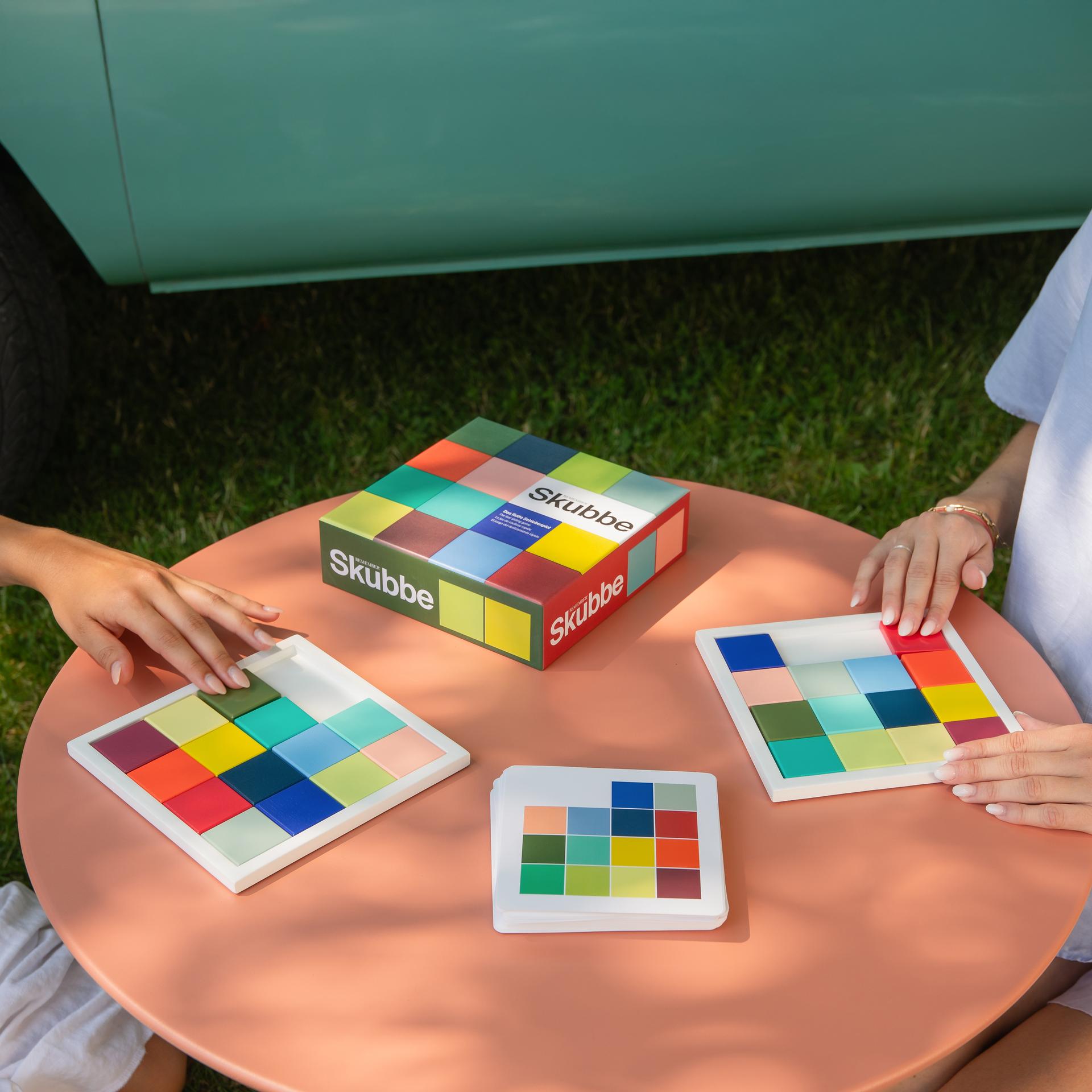 Zwei Personen spielen Skubbe am Tisch. Das Spielbrett, Karten und die Verpackung sind zu sehen. Bunte Spielsteine liegen auf den Brettern.