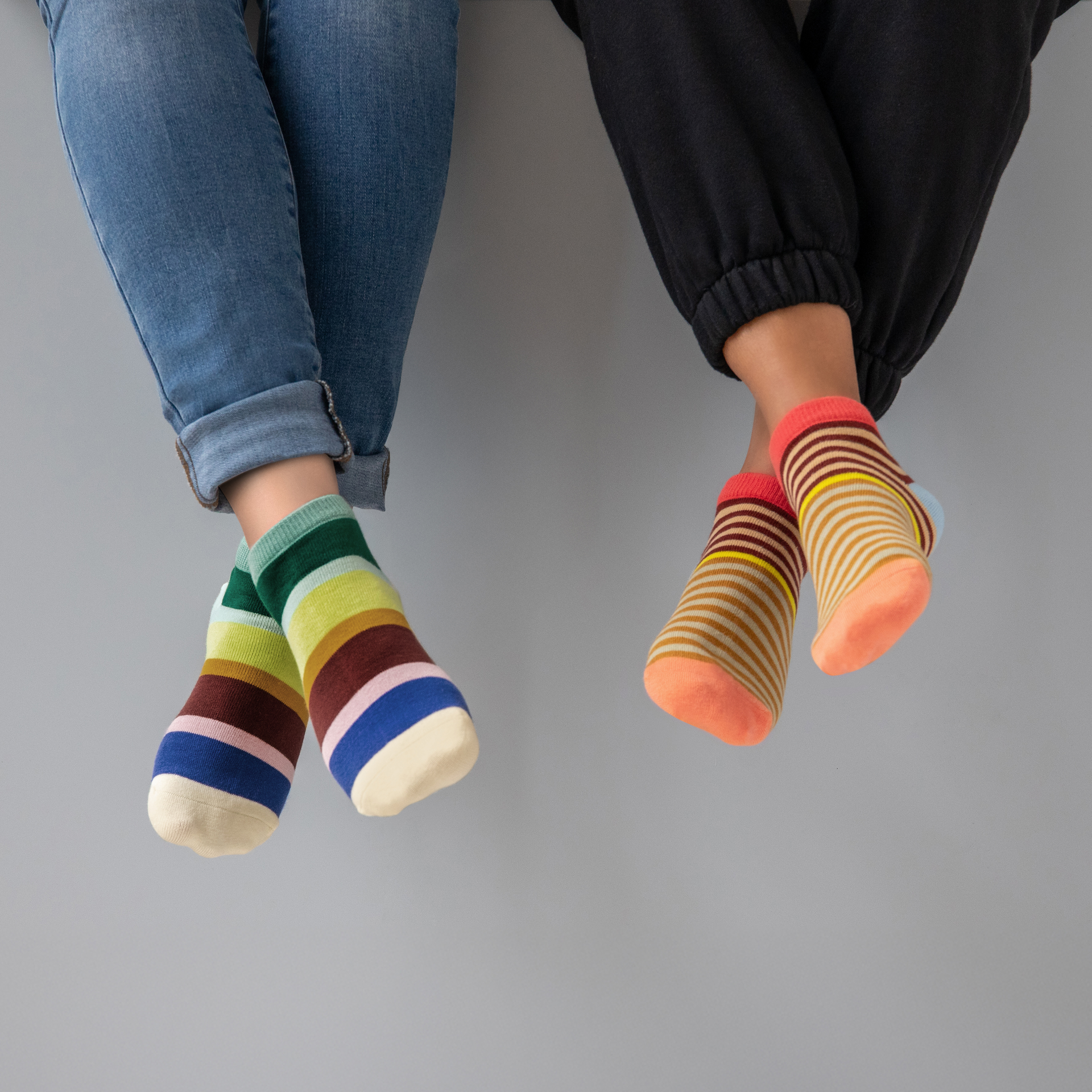 Zwei Paar Füße mit bunten gestreiften Sneakersocken der Größe 36-41. Ein Paar in Jeans, das andere in Jogginghose.