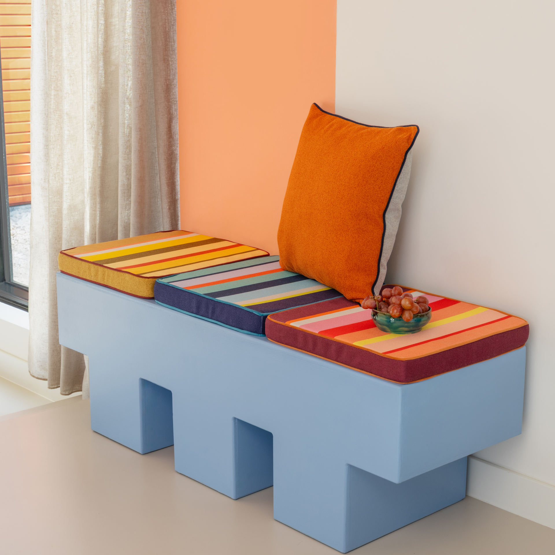 Einrichtungsecke mit hellblauem Podest, farbigen Sitzkissen und orangefarbenem Kissen. Eine Schale mit Weintrauben steht auf einem der Kissen. Im Hintergrund eine orange Wand.