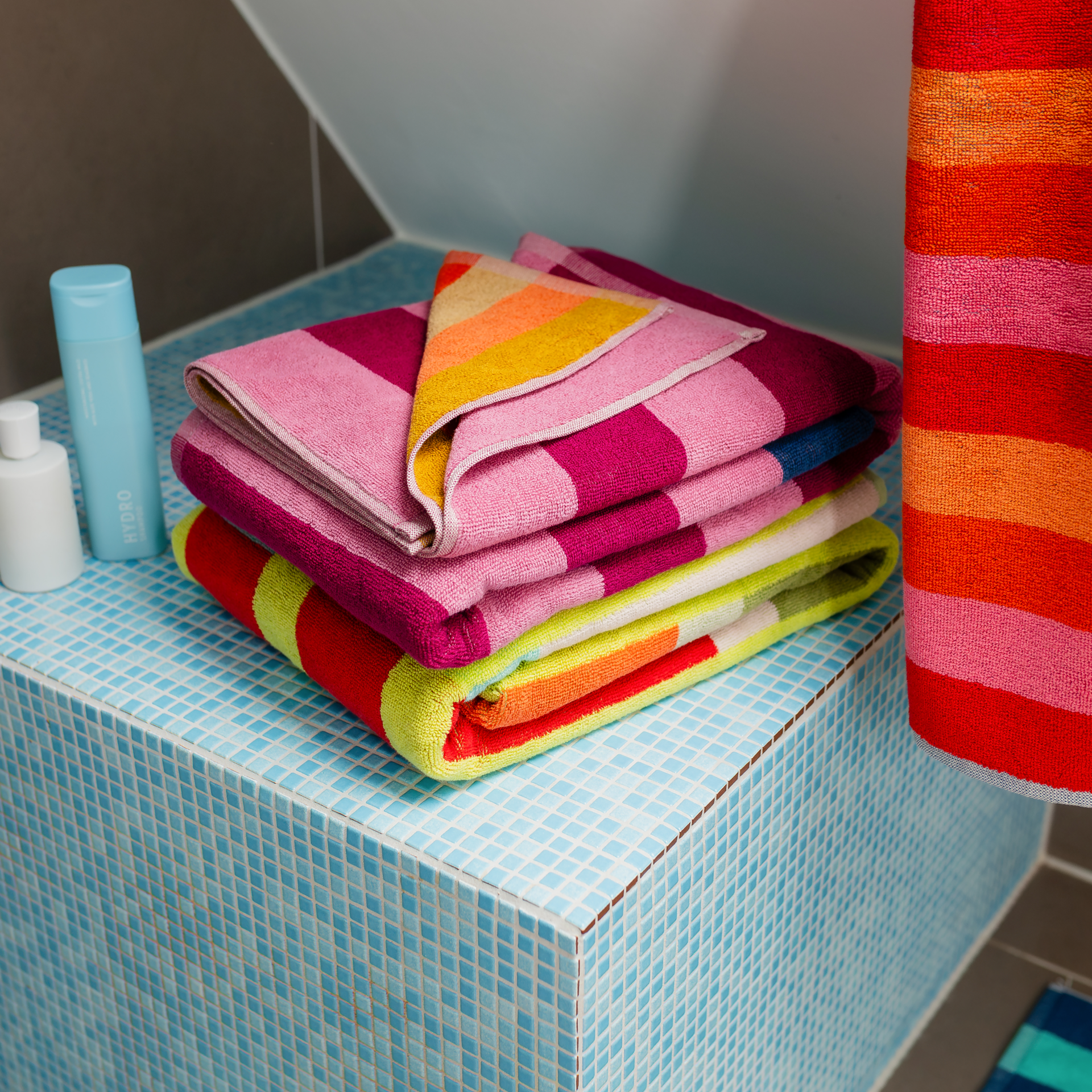 Serviette de bain Alva pliée verticalement, avec des rayures transversales colorées : en haut, alternance de violet foncé et violet clair, puis bleu foncé et turquoise clair ; en bas, alternance d'orange et de beige. Très grande, moelleuse, douce.