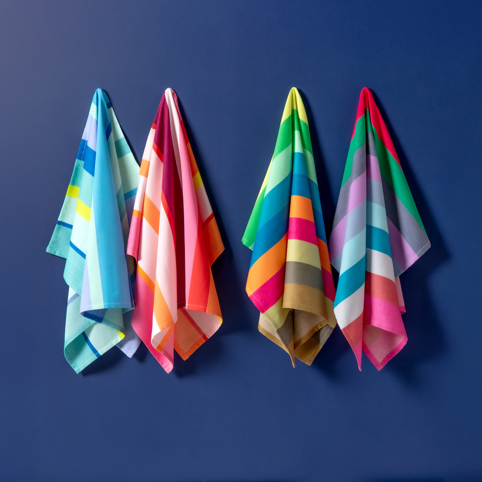 Vier bunte Tücher sind an einer blauen Wand platziert. Sie zeigen Streifen- und Karomuster in den Farben blau, grün, gelb, rot und pink. Die Tücher sind variantenreich drapiert.