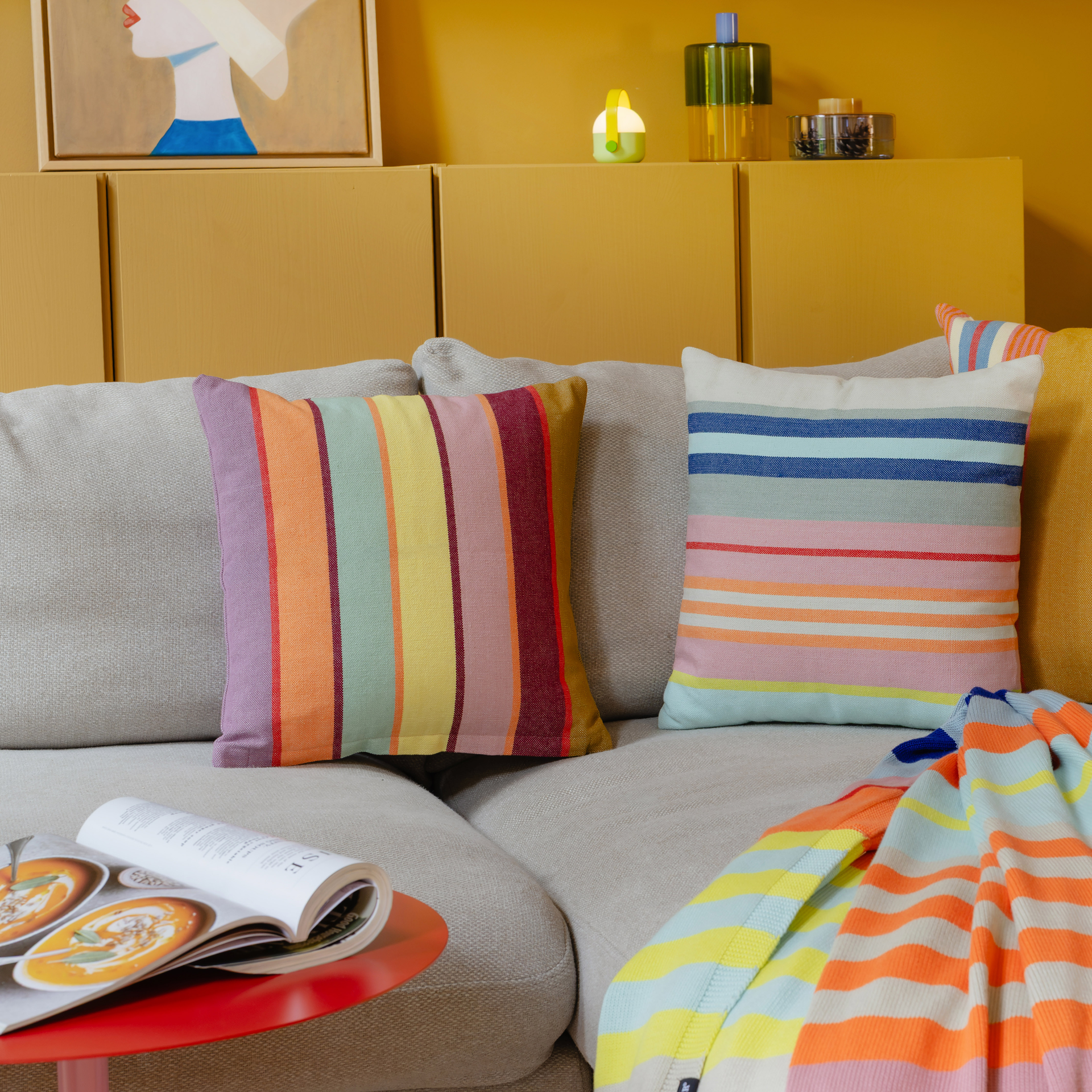 Cuscino quadrato in cotone con vivaci strisce verticali in diversi colori: viola, arancione, verde, giallo, rosa, bordeaux e verde oliva. Struttura a trama ben definita e piccola etichetta del marchio nera in basso a destra.