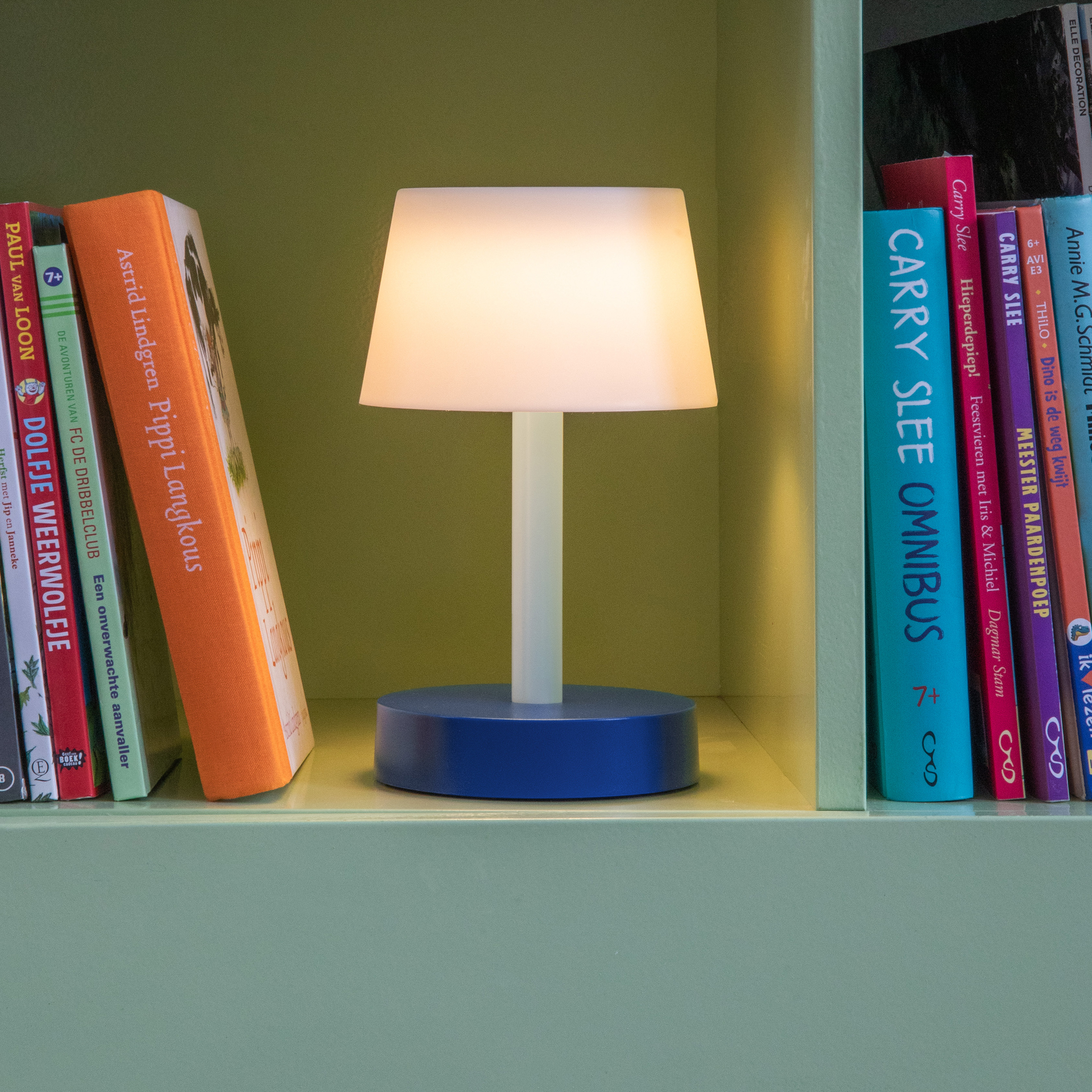 Ambiente-Aufnahme der Tischleuchte Fritz 'Sky' mit blauem Sockel und leuchtendem, weißem Schirm im Regal zwischen Büchern.