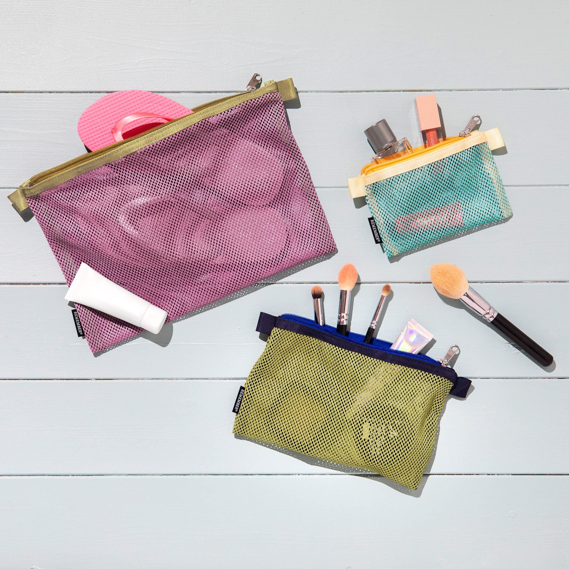 Drei farbige Mesh Bag 'Jordy' Sets, gefüllt mit Kosmetika, auf hellblauem Holzuntergrund. Die Taschen sind in pink, türkis und olivgrün.