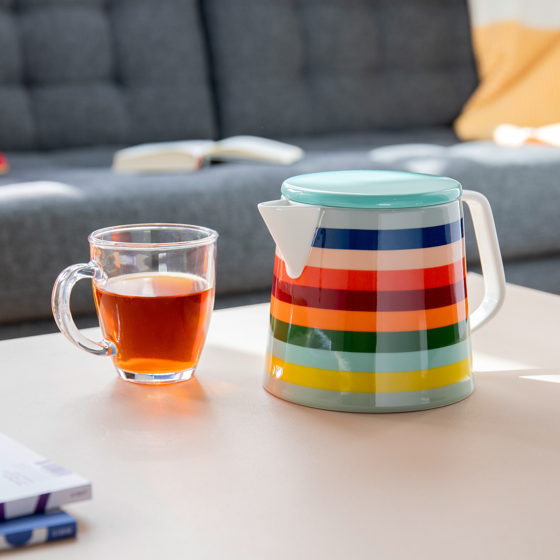Eine Teekanne 'Lorenzo' mit bunten Streifen steht neben einer Tasse Tee auf einem Tisch. Im Hintergrund ist ein graues Sofa zu sehen.