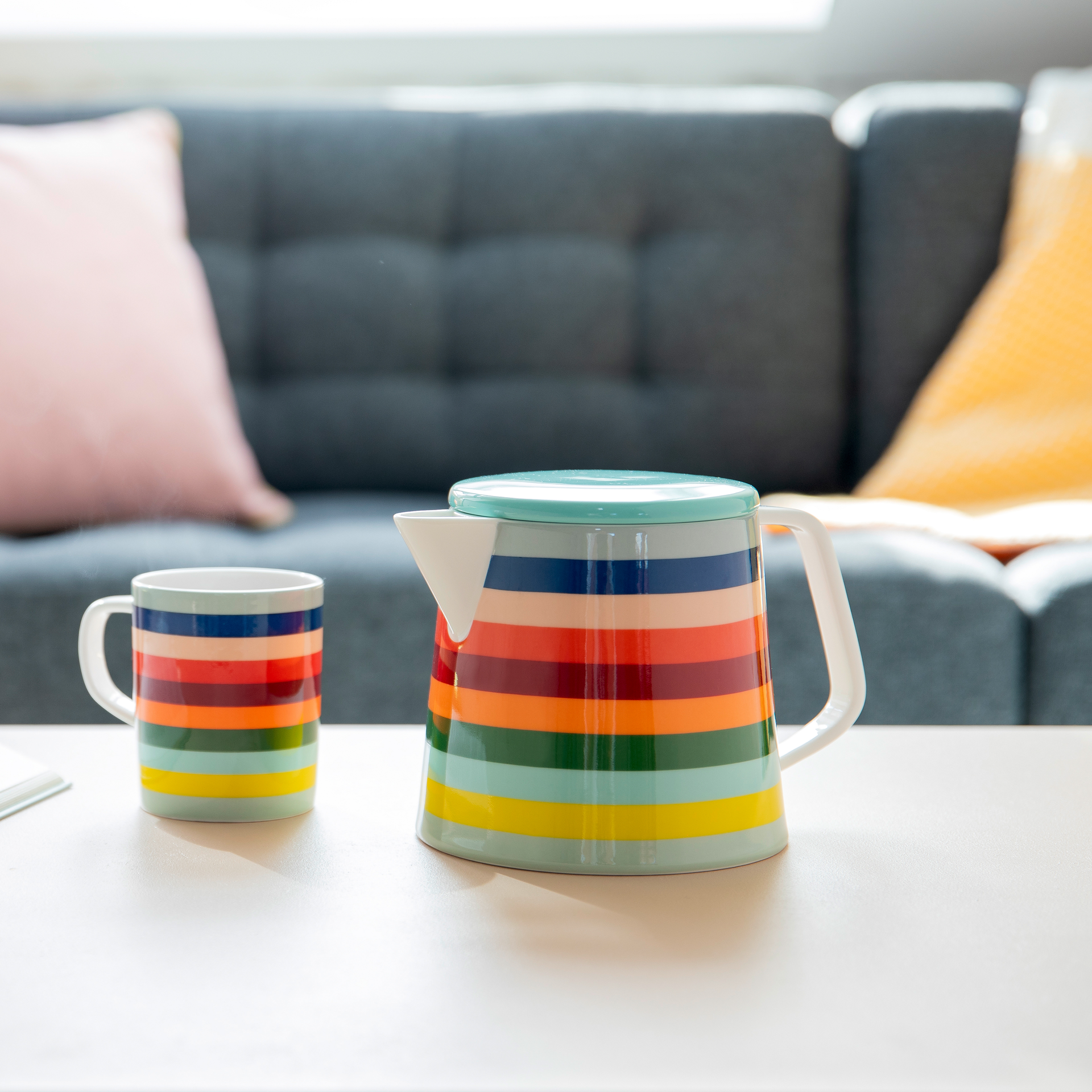 Eine 'Lorenzo' Teekanne und eine passende Tasse mit bunten, horizontalen Streifen stehen auf einem Tisch. Im Hintergrund ist ein graues Sofa zu sehen.