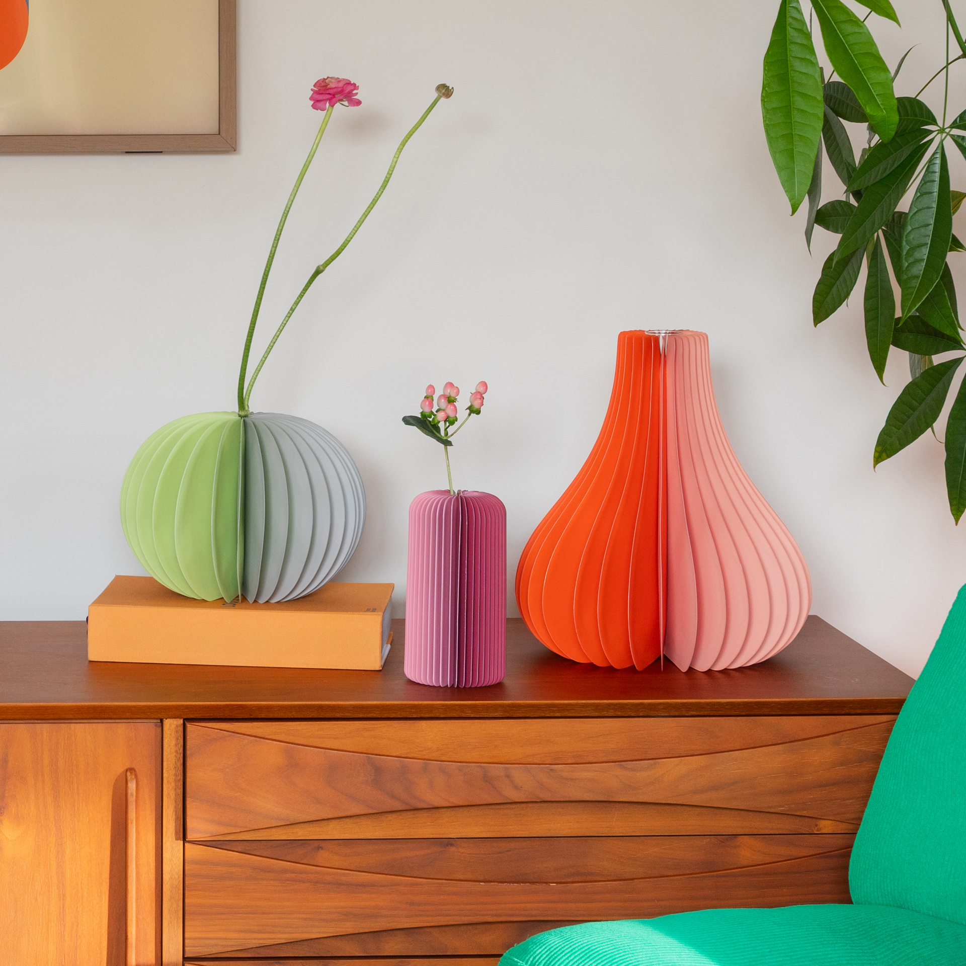 Trois vases en papier orange, rose, vert et gris, garnis de fleurs, posés sur une commode.