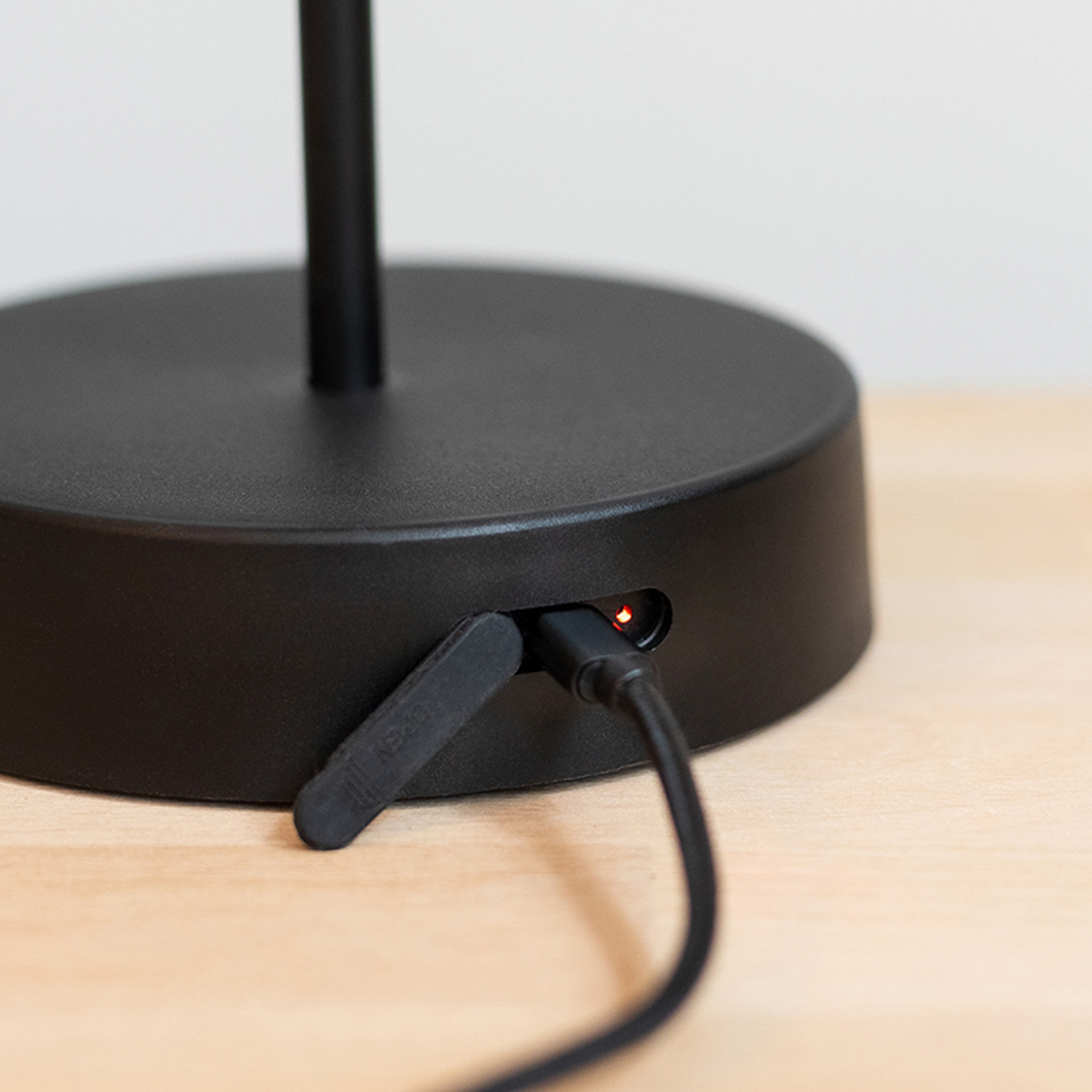 Detailaufnahme der Tischleuchte Oscar im 'pure' Design mit eingestecktem USB-Kabel und roter Ladeanzeige am Sockel auf einem hellen Tisch.