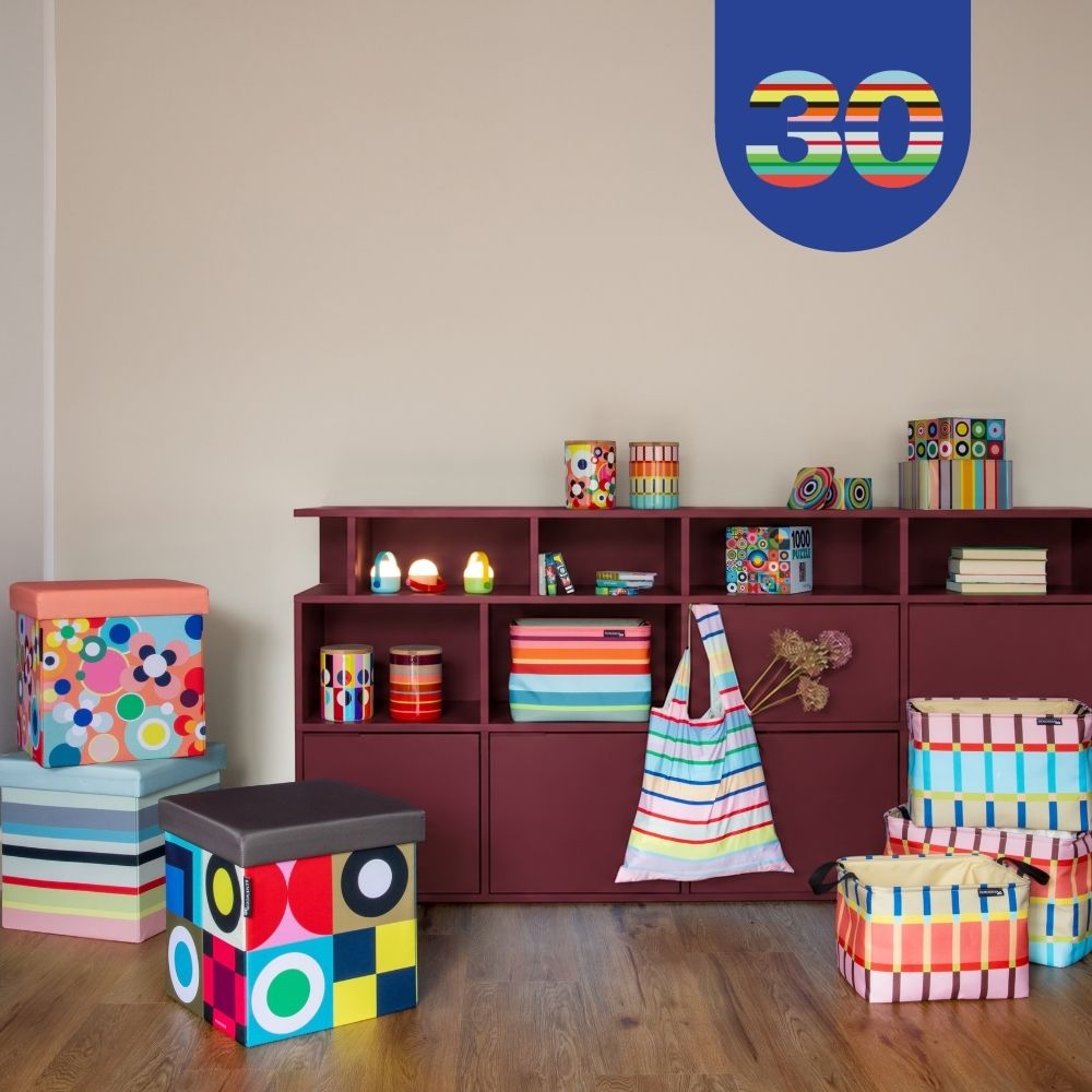 Pièce colorée avec une étagère bordeaux, remplie de boîtes de rangement multicolores et d’objets décoratifs. Sur le parquet, on trouve des tabourets et des paniers à motifs. En haut à droite sur le mur, un logo bleu avec le chiffre « 30 ».