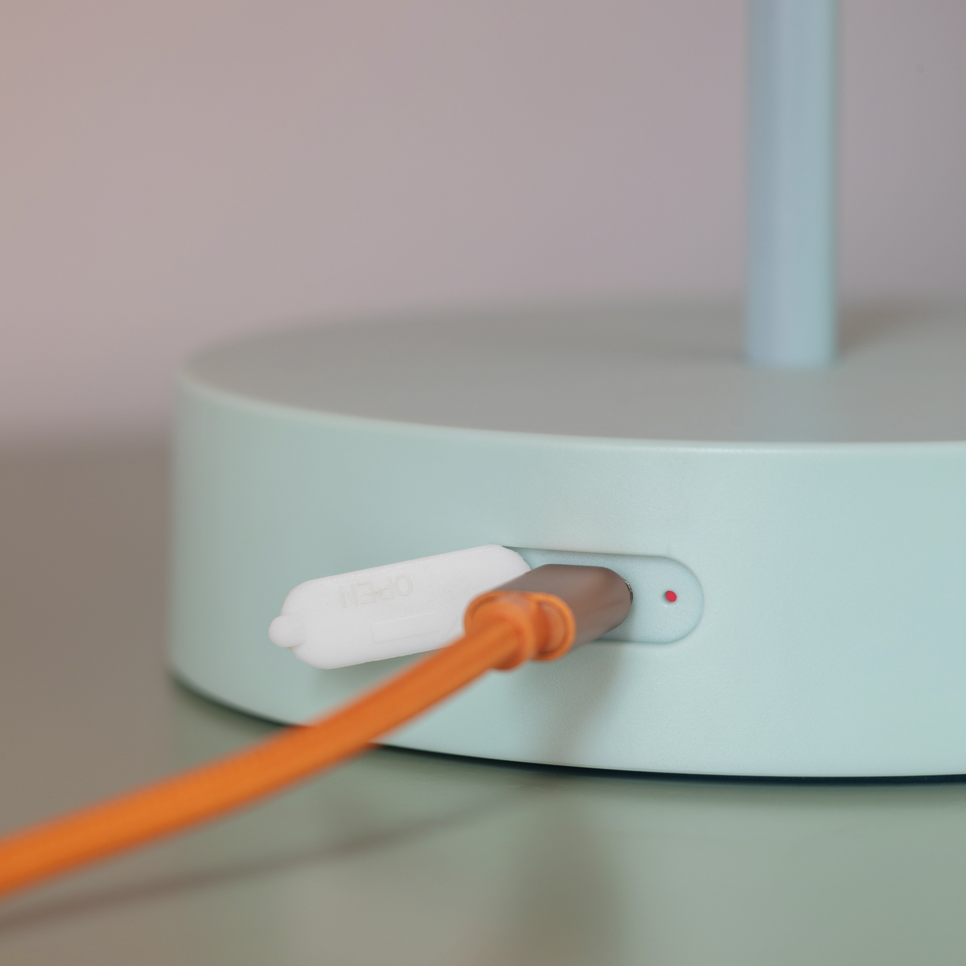 Zoom auf die Tischleuchte Oscar: Der orangefarbene USB-Stecker ist in die mintgrüne Leuchtenbasis eingesteckt. Daneben befindet sich eine USB-Buchse mit einer weißen Abdeckkappe. Ein roter Punkt ist sichtbar.