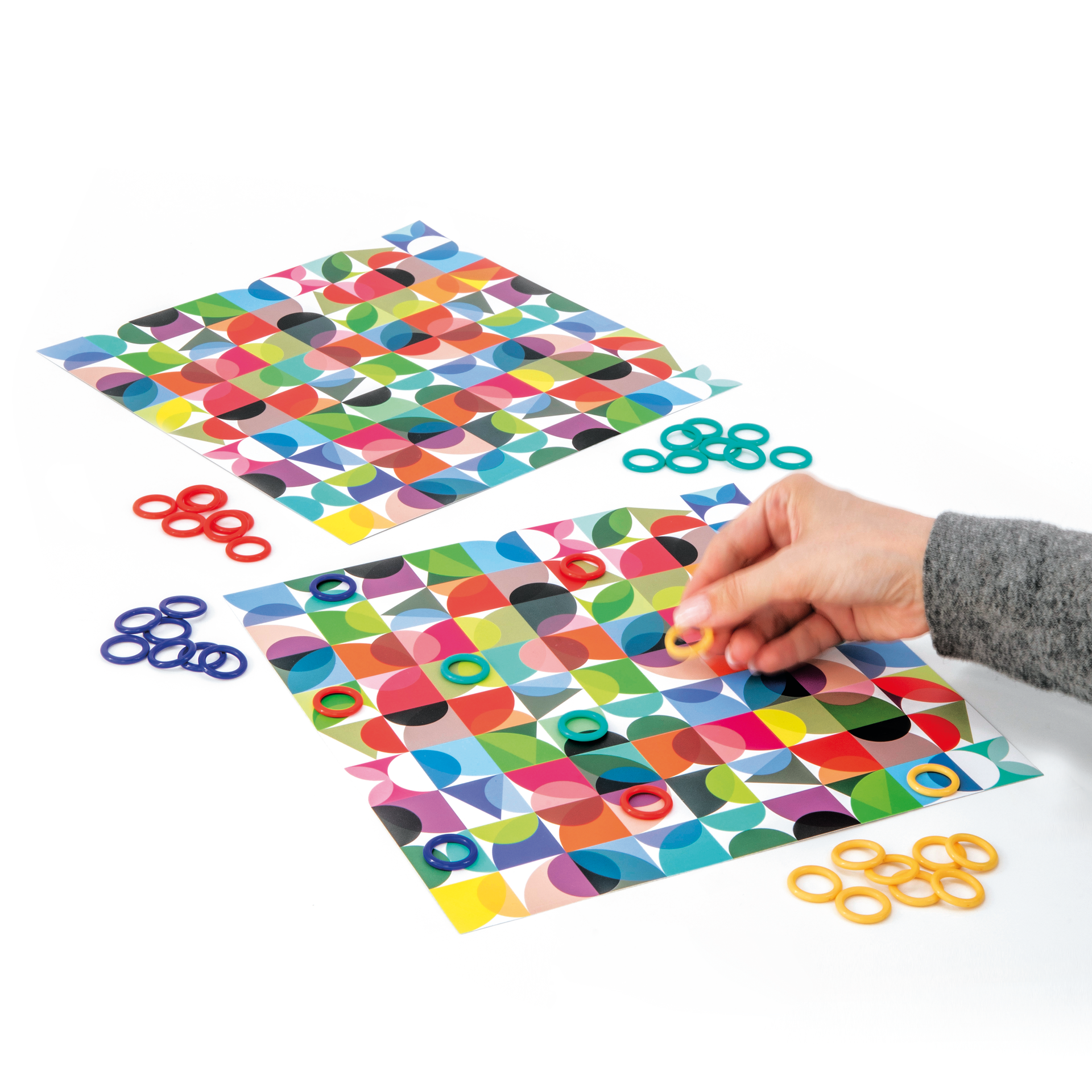 Zwei Double Spielblätter mit bunten geometrischen Mustern liegen im Warenkorb. Farbige Ringe markieren Fehler. Eine Hand legt einen gelben Ring.