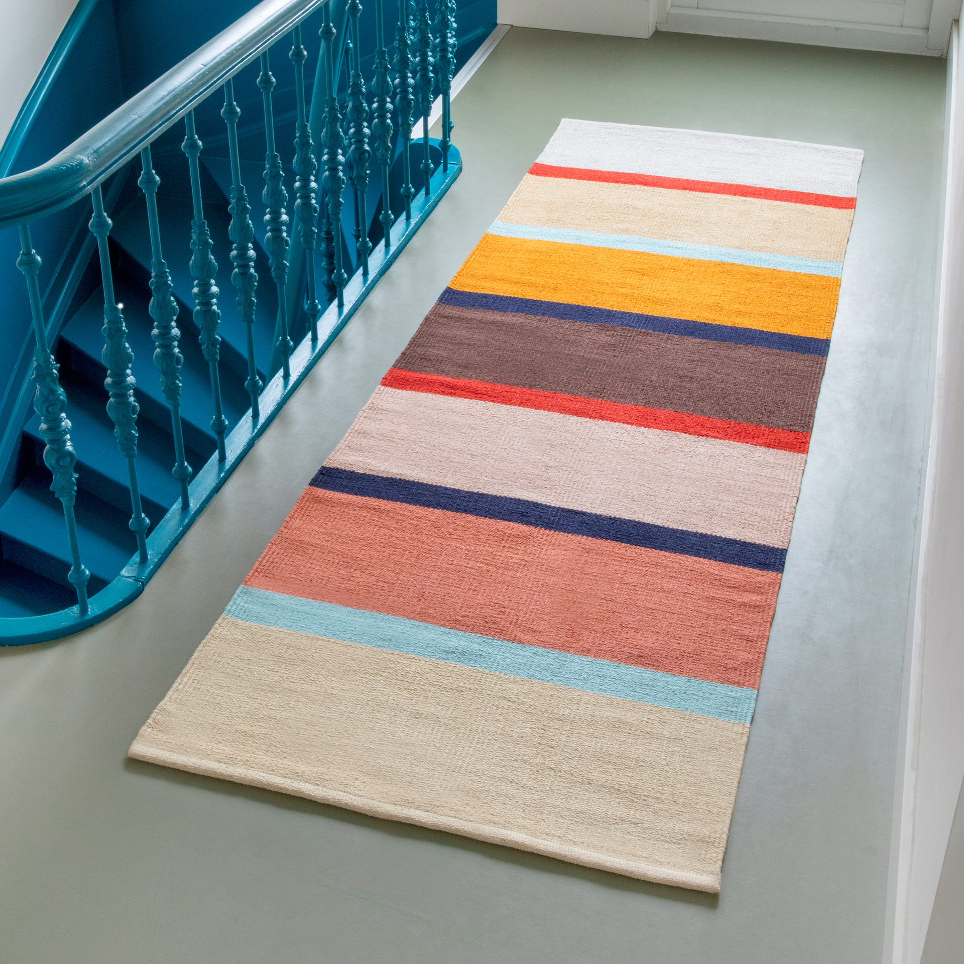 Tapis de sol 'Zaza' tissé à la main avec des rayures beiges, bleues, orange, marron et rouges sur un sol gris à côté d'un escalier bleu.