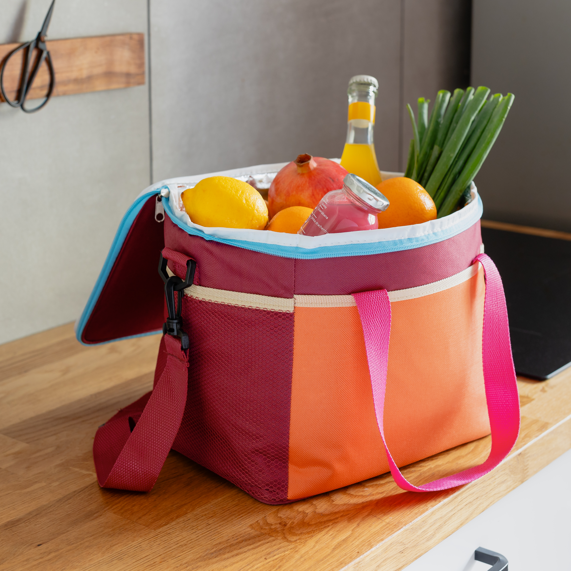 Freistehende, bunte Kühltasche 'Telli' mit Trageriemen und Schultergurt. Der obere Teil ist rot mit einem hellblauen Reißverschluss. Die Mitte ist orange, die Seite rot-netzartig.
