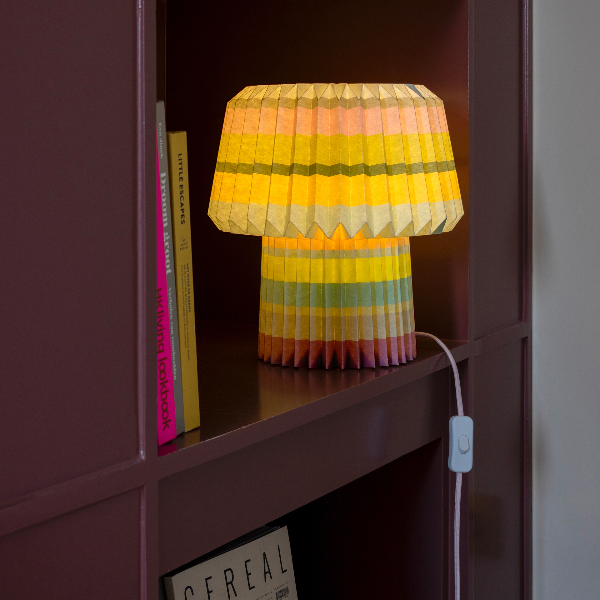 Das Produkt Papierleuchte Anna 'stripes' steht auf einem Regal und leuchtet. Der Lampenschirm hat ein gestreiftes Muster in Gelb-, Rosa- und Grüntönen. Bücher stehen daneben.